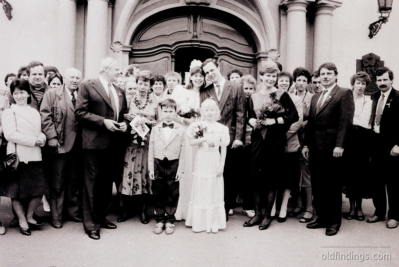 A black-and-white wedding photo featuring a bride in a classic A-line gown and groom in a suit, flanked by 16 guests in 1970s attire. The couple stands outside a grand, arched entrance with ornate stonework, likely a church or civic building. Guests pose formally, holding bouquets or programs.