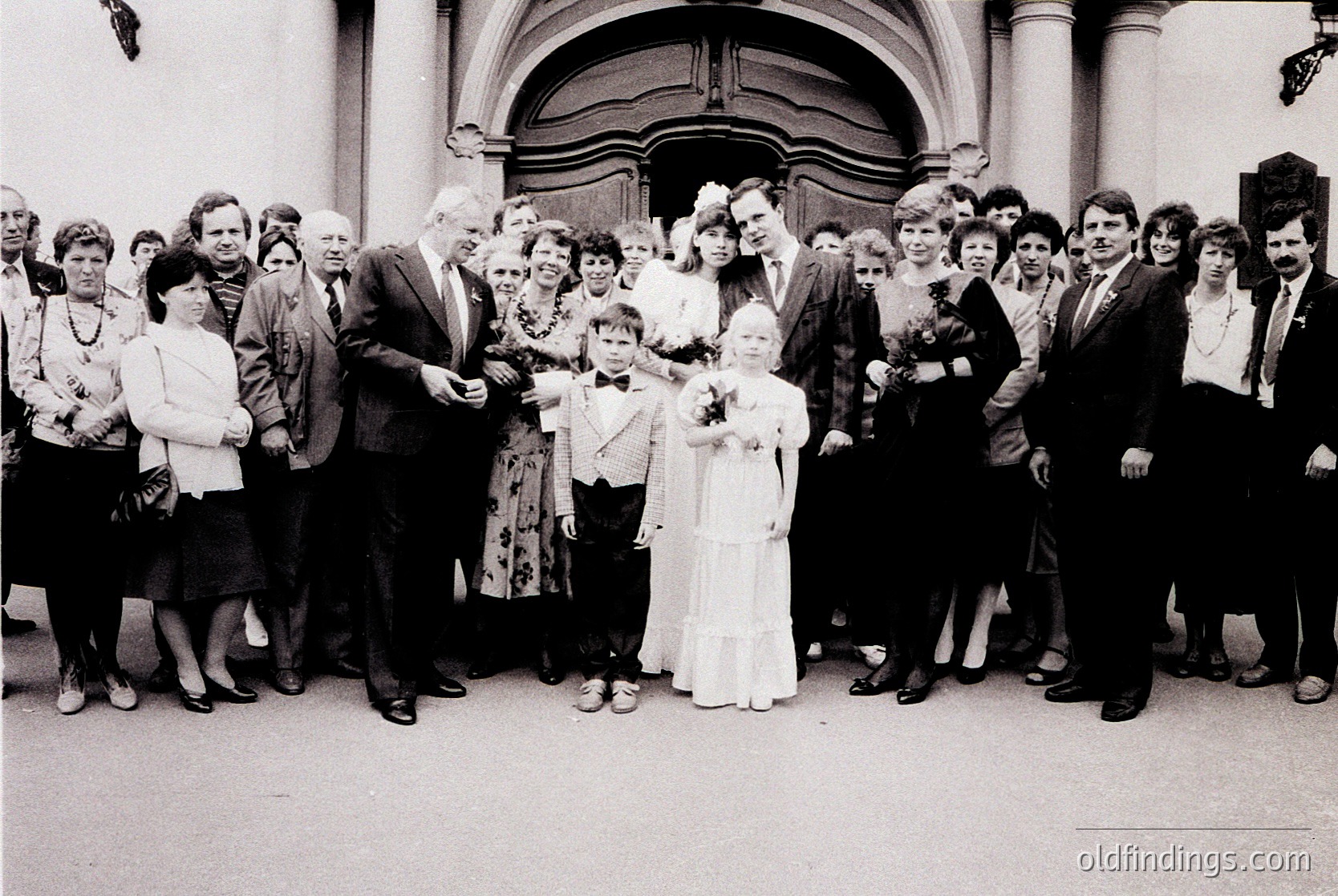 Family wedding photo outside a grand, arched entrance, likely 1970s–1980s. Bride in white gown with bouquet, groom in suit, surrounded by 16 guests in formal attire. Classic black-and-white composition with vintage clothing and hairstyles.