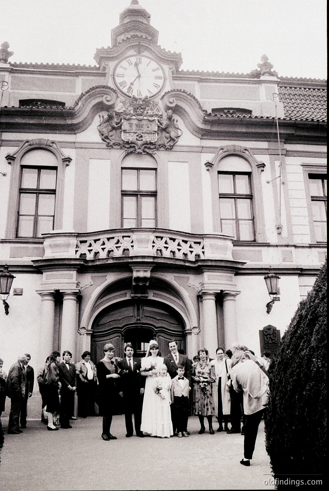 Neoclassical building facade featuring a prominent clock tower and arched entrance, flanked by columns. A wedding party stands in the courtyard, with the bride in a white gown and groom in a suit. Mid-20th century attire suggests or . Architectural details include ornate moldings and symmetrical windows.