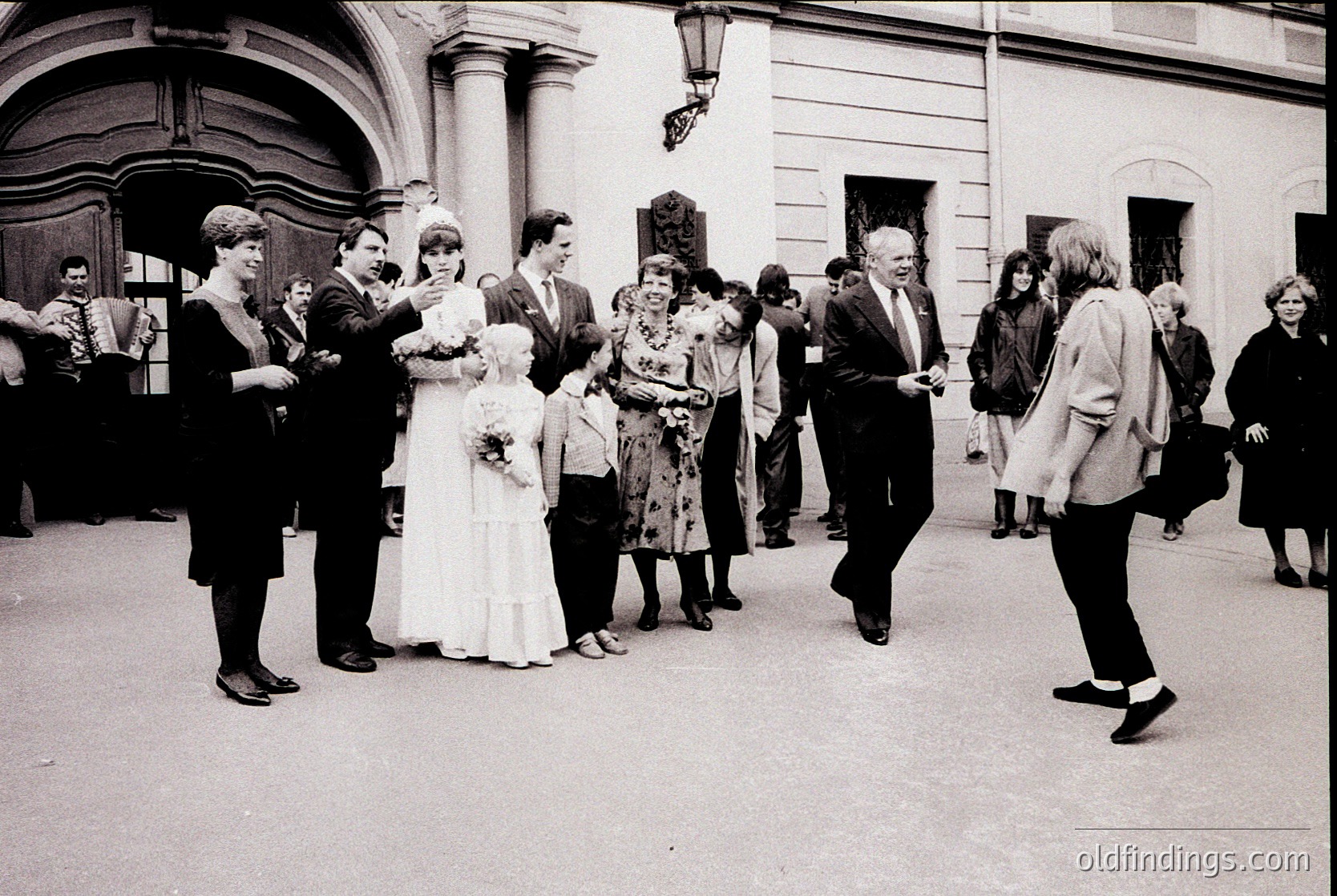 Neoclassical courtyard wedding celebration, 1960s-1970s. Bride in full-length white gown with floral bouquet, surrounded by guests in formal attire—men in suits, women in dresses. Architectural details include tall columns and arched doorways. Candid moments capture joyful interactions.