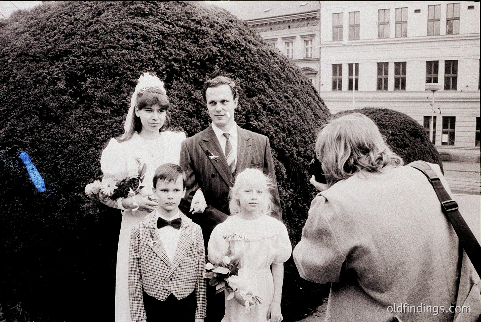 A 1960s-70s family portrait in urban setting: bride in lace gown, groom in suit with bow tie, two children in formal attire holding flowers. Photographer captures candid moment from behind. Architectural backdrop: multi-story building with classical details.