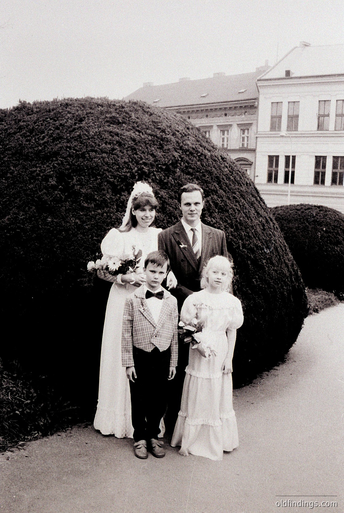 Family portrait featuring a bride in a full-length white gown with floral bouquet, groom in a dark suit, and two children in early 1960s attire. Greenery and classical European architecture in background. Candid, outdoor setting.