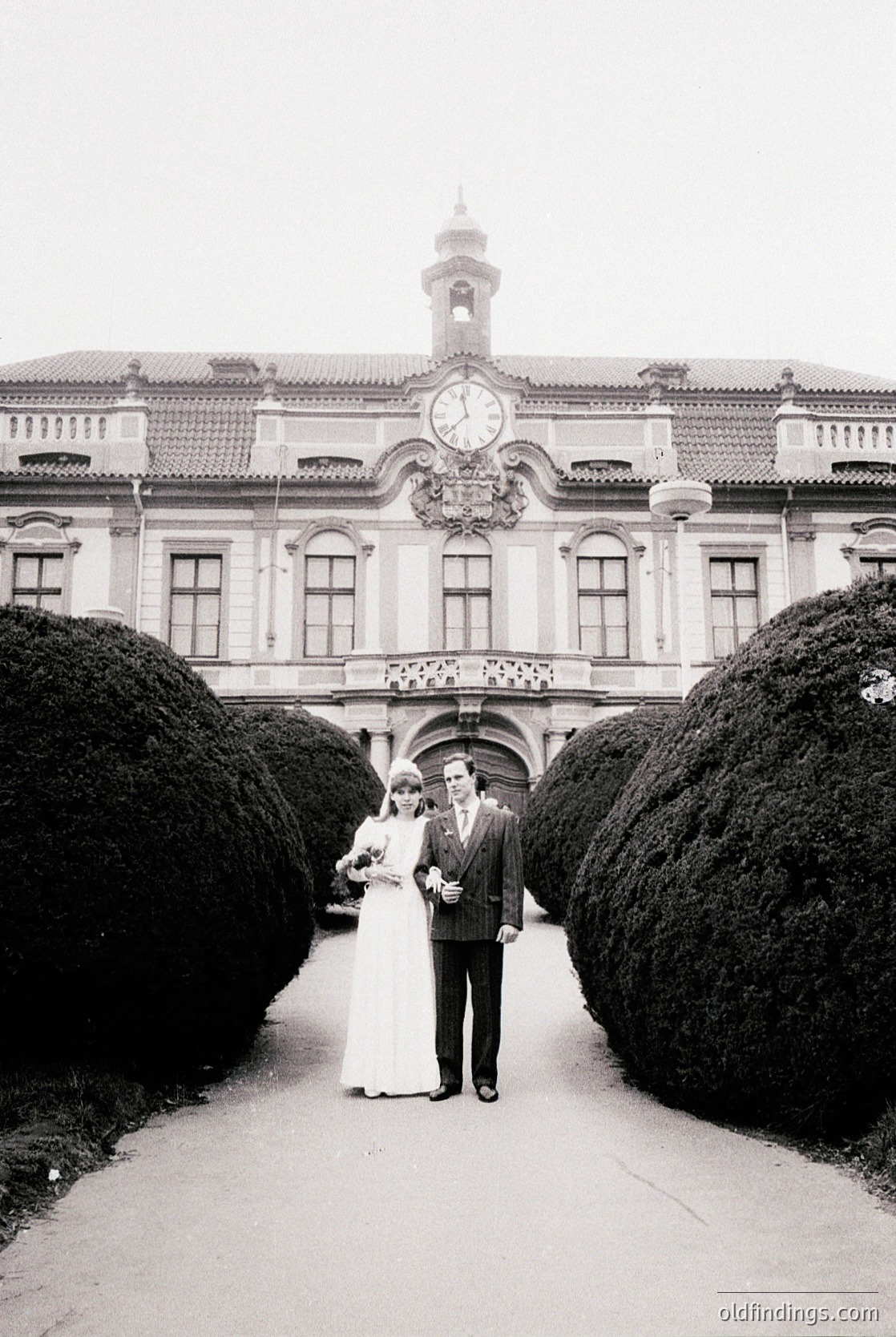 Classic 1960s-70s wedding portrait in grand European courtyard. Couple in formal attire—bride in floor-length gown, groom in suit—posing under ornate clock tower with Baroque architectural details. Manicured topiary hedges frame the scene. Timeless elegance with vintage charm.