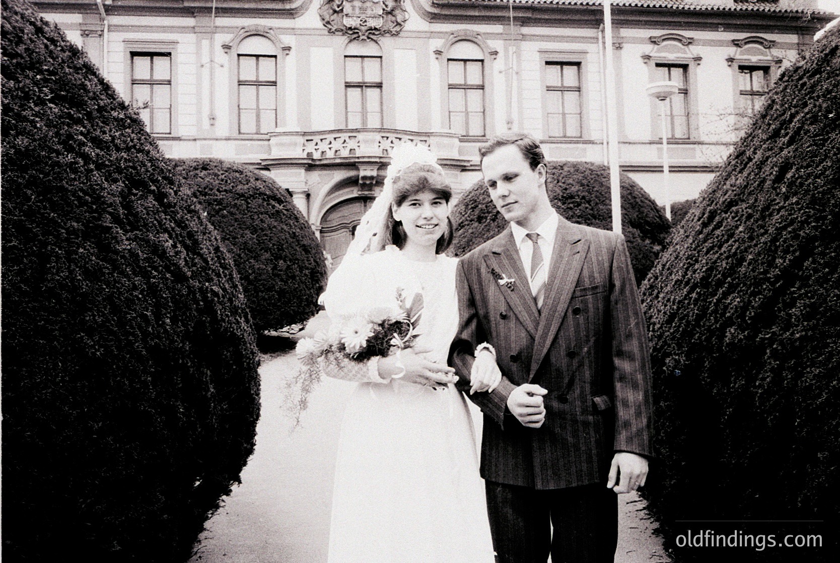1970s black-and-white wedding portrait in European classical courtyard. Bride in full-length gown with bouquet, groom in pinstripe suit. Ornate stone façade with arched windows and trimmed hedges. Timeless formal attire and architecture evoke mid-century European elegance.