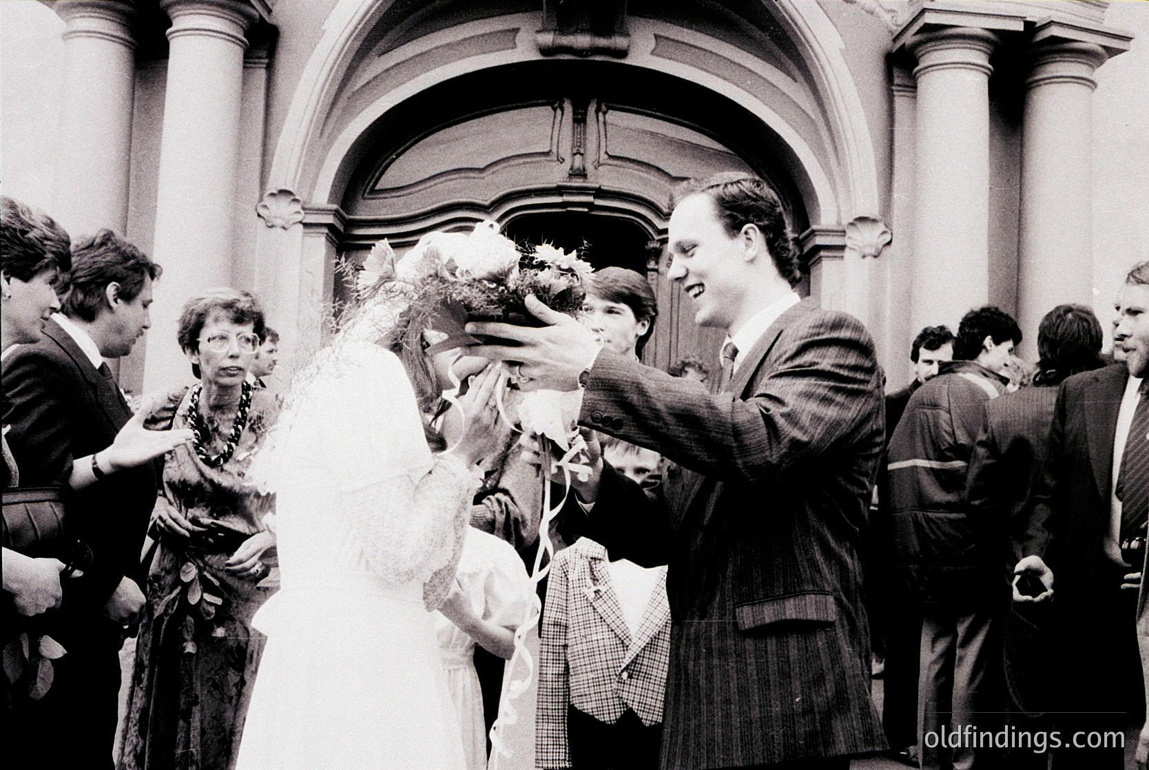 Mid-20th century wedding scene outside grand neoclassical building. Bride in traditional veil and groom in suit toss confetti to cheering crowd. Formal attire and architectural details suggest European setting, likely 1950s–1960s.