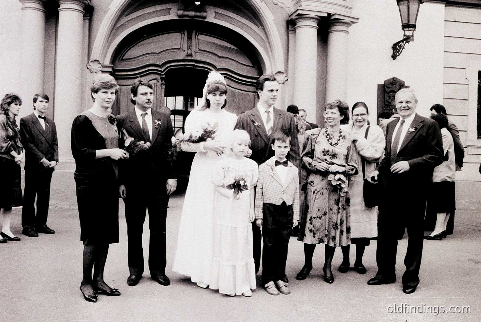 Neatly posed wedding group photo in front of a grand, arched entrance with classical columns. Bride in simple white gown with bouquet, flanked by family in 1970s-era attire—men in suits, women in floral dresses. Candid expressions and formal poses blend. Likely European setting.