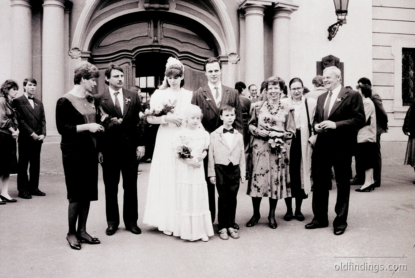Neoclassical building entrance with arched doorway, featuring a 1970s wedding group portrait. Bride in full-length gown with bouquet, flanked by groom in suit and two children in formal attire. Guests in vintage 1970s clothing—men in suits, women in floral dresses. Candid, outdoor setting with street lamp and stone steps.