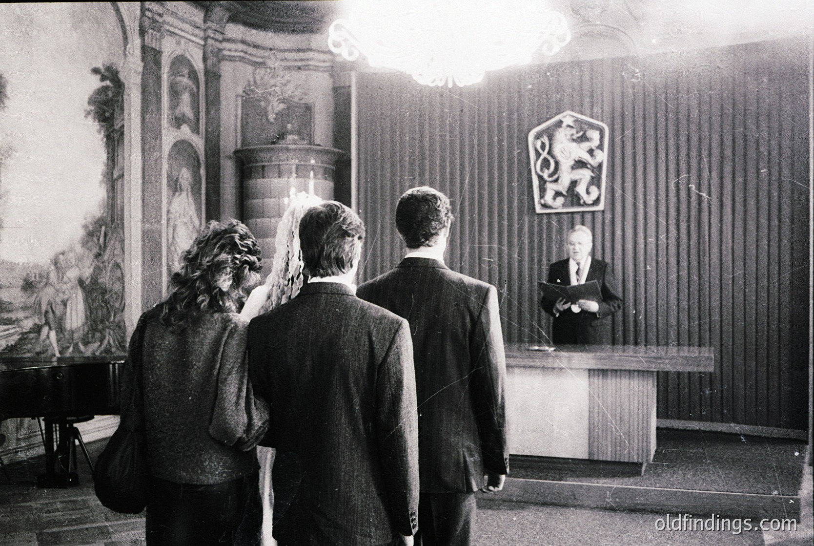 Black-and-white formal event in ornate hall featuring three men in suits and a woman in a veil, facing a podium with a coat-of-arms emblem. Baroque-style frescoes and columns enhance the setting, suggesting a 1960s–1970s European ceremony.
