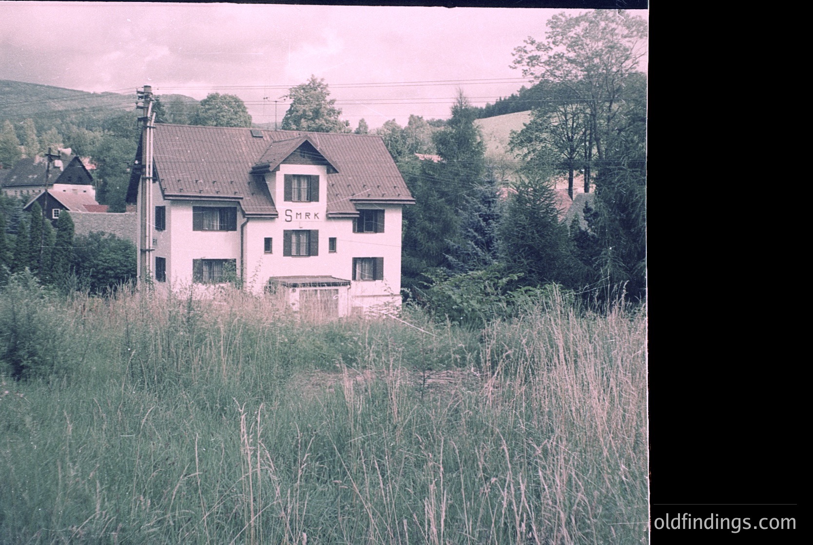 Two-story alpine-style building with "Snax" signage, surrounded by overgrown grass and forested hills. Likely mid-20th century European mountain retreat or guesthouse.