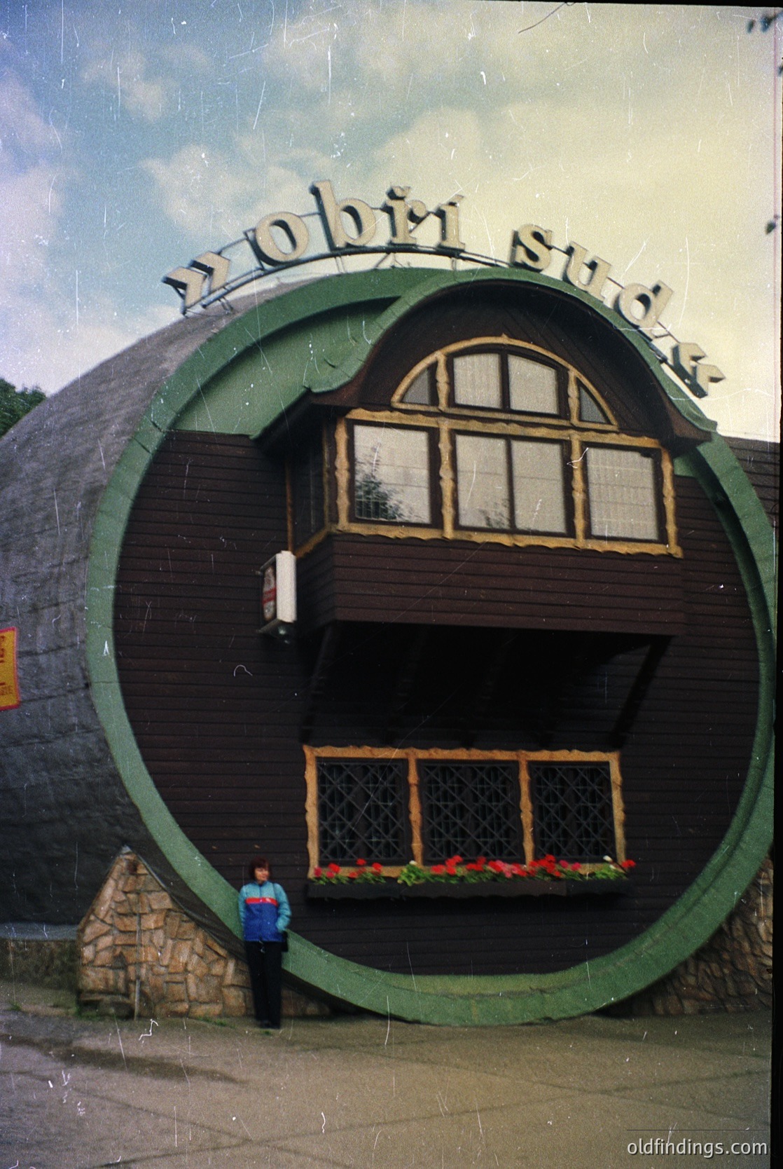 Retro circular building with "Pobit sud" signage, likely a 1970s-era public facility in Bulgaria. Wooden facade with arched windows and red flowers below. Circular entrance framed by stone pillars. Vintage photo quality with slight grain.