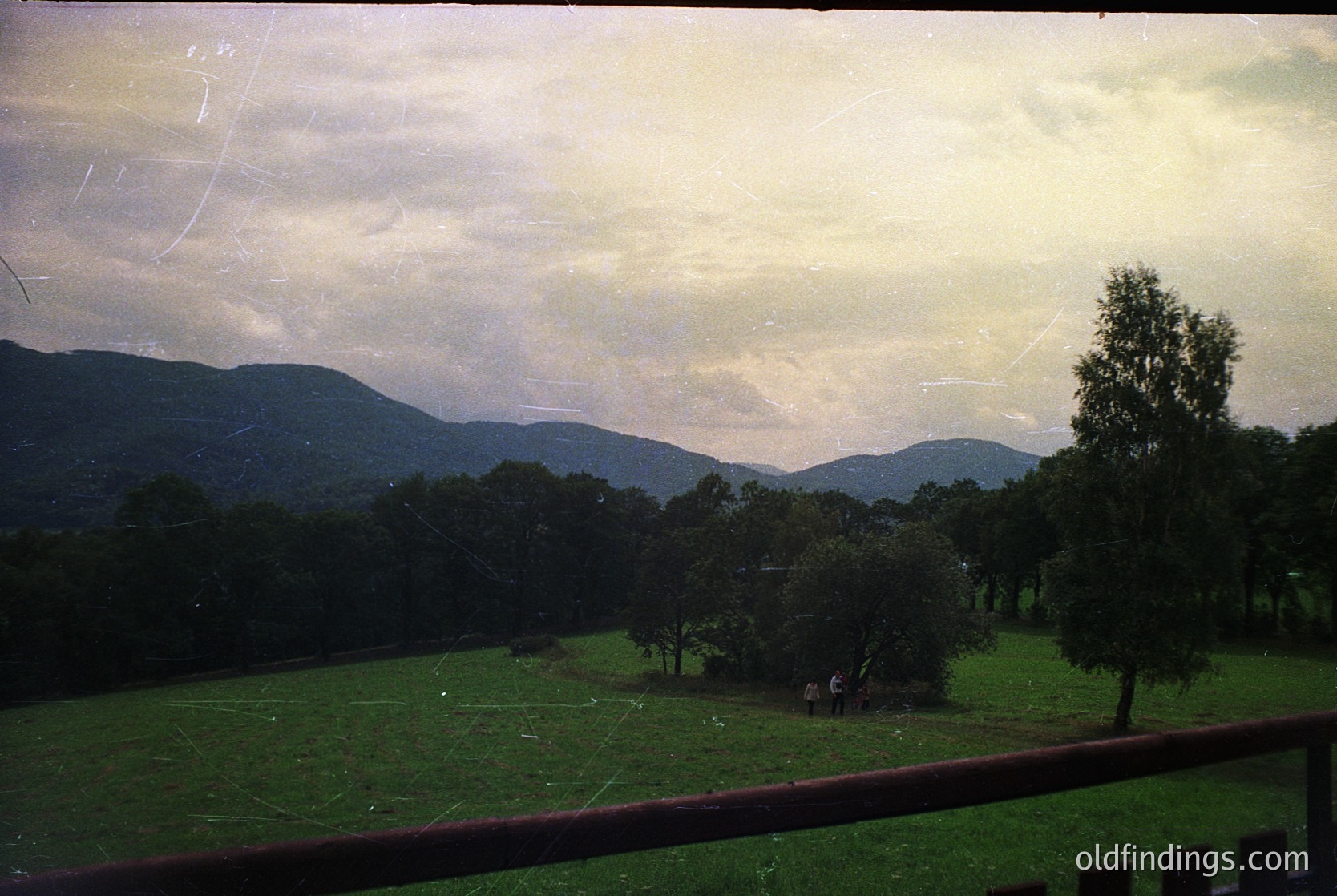 Vintage landscape shot through a window frame, showcasing rolling hills and dense forest under overcast skies. Two figures stand near a small cluster of trees in a lush green meadow. Likely Eastern European countryside, 1970s–1980s era.