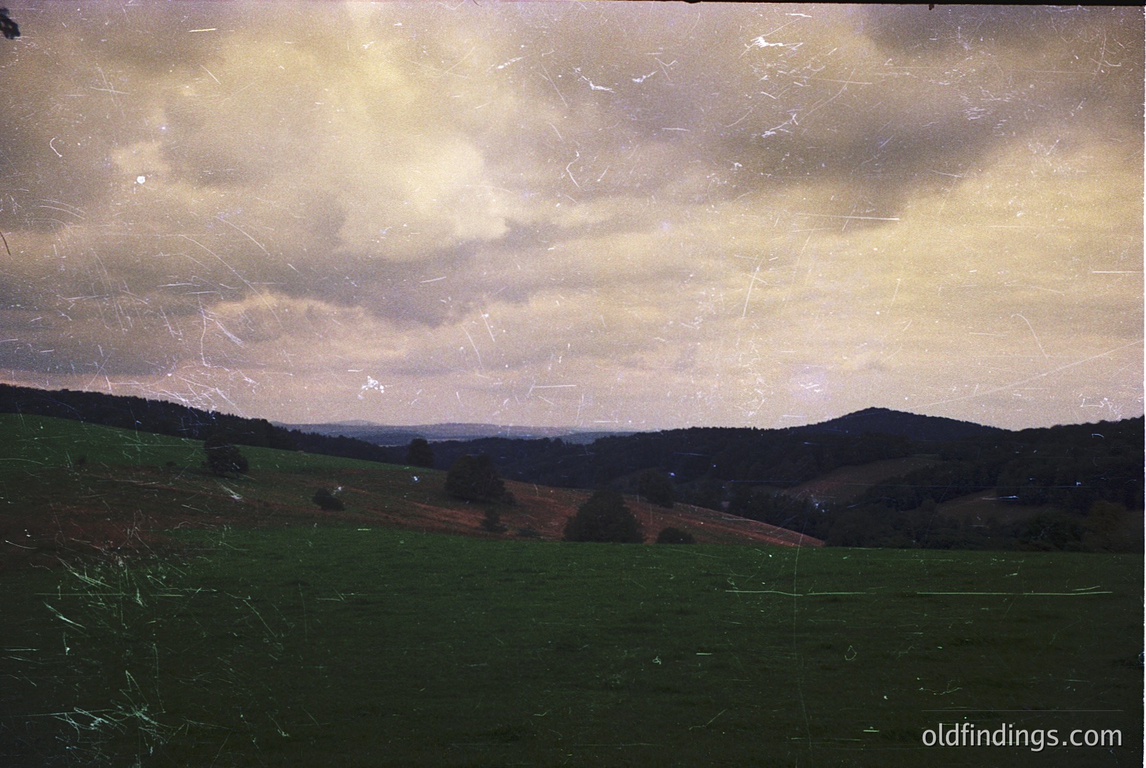 Vintage landscape photograph featuring rolling hills under dramatic, textured clouds. Evidence of aging film grain and slight scratches. Likely captured during golden hour for soft lighting.