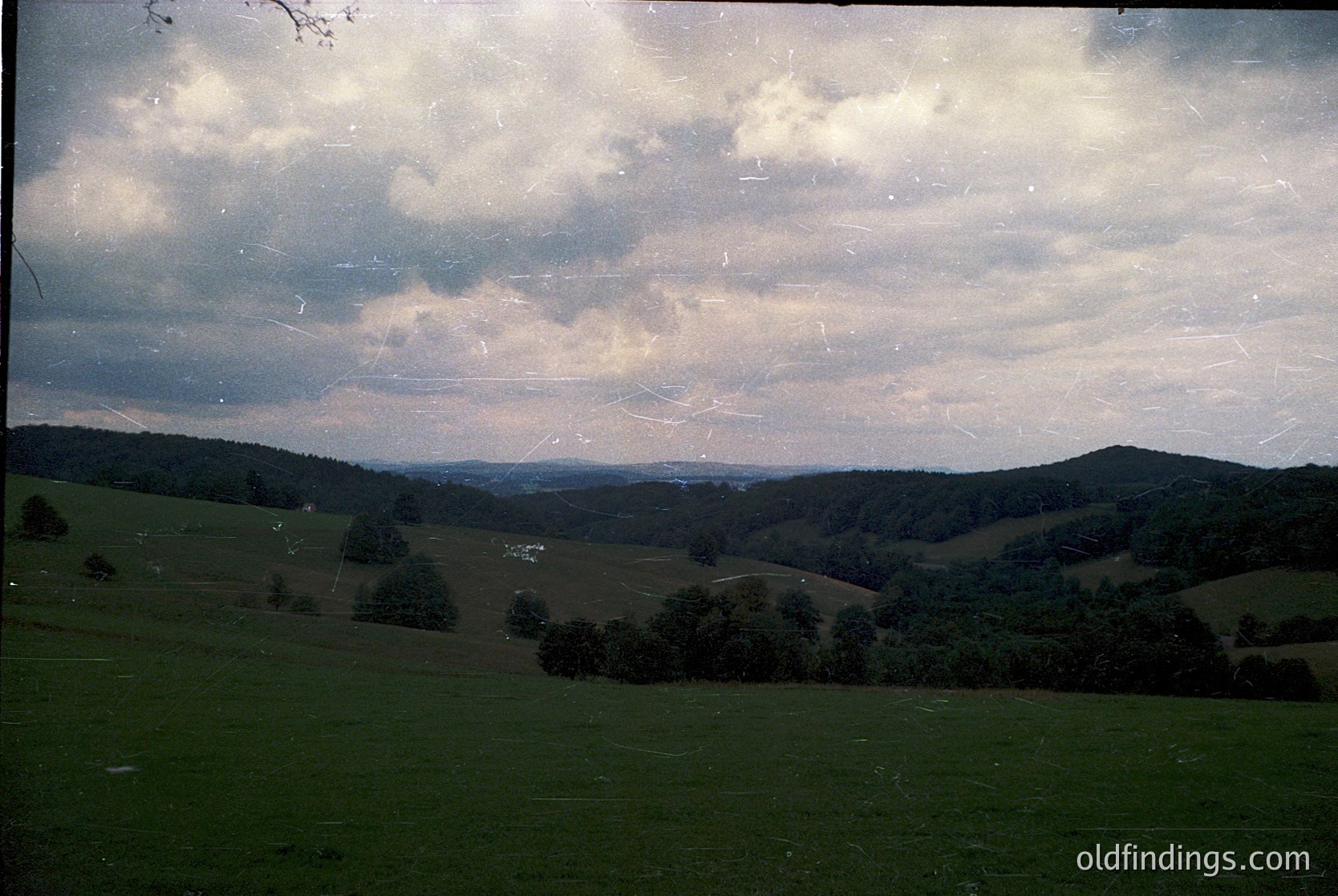 Vintage landscape shot of rolling hills under dramatic cloud cover, likely mid-20th century. Green pastures with scattered trees frame distant ridges, suggesting rural Europe. Film grain and slight yellow tint indicate analog photography. Ideal for historical research or nostalgic design references.