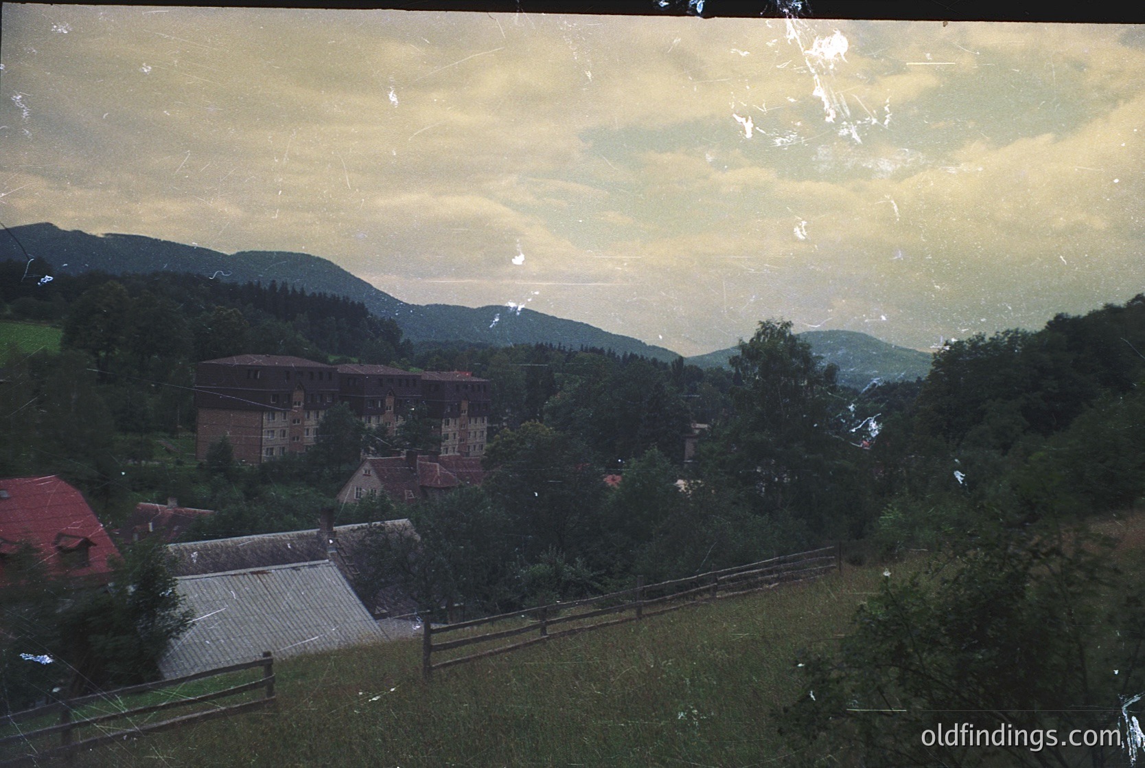 Vintage landscape shot featuring a rustic village nestled in hilly terrain. Mid-20th century architecture with brick buildings and red-tiled roofs. Lush greenery and wooden fences frame the foreground. Overcast sky enhances muted tones. Likely Eastern European countryside.