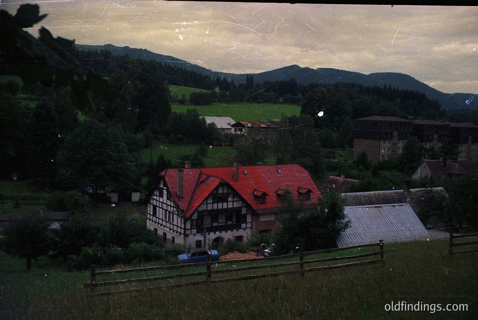 Vintage sepia-toned photo of a half-timbered chalet-style house with red roof, nestled in a lush valley surrounded by forested hills. The structure features exposed wooden beams and a small porch. Likely European alpine region, mid-20th century.