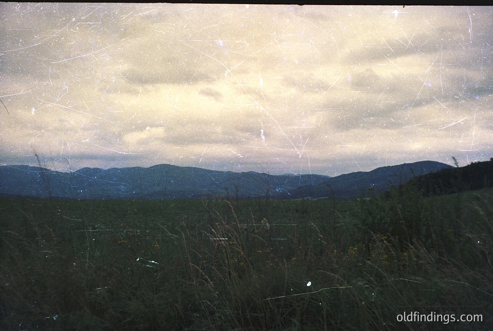 Vintage sepia-toned landscape with cracked, aged film texture. Rolling hills and open meadows under dramatic, cloud-laden sky. Likely Eastern European countryside, 1960s–1980s.