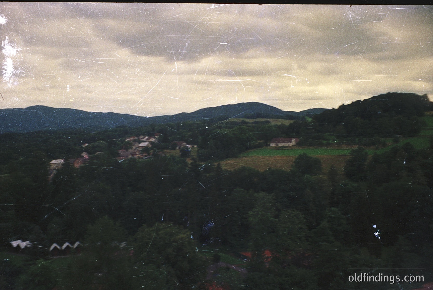 Vintage aerial view of rural landscape with dense forest, scattered farmhouses, and rolling hills under overcast skies. Scratched film grain suggests mid-20th century photography.