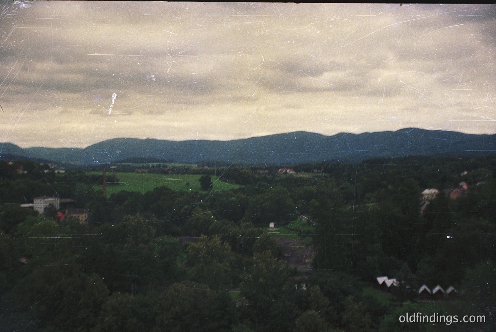 Vintage aerial view of rural landscape with dense forest, rolling hills, and scattered buildings. Overcast sky with visible scratches and fading edges suggests aged film. Likely Eastern European countryside, mid-20th century.