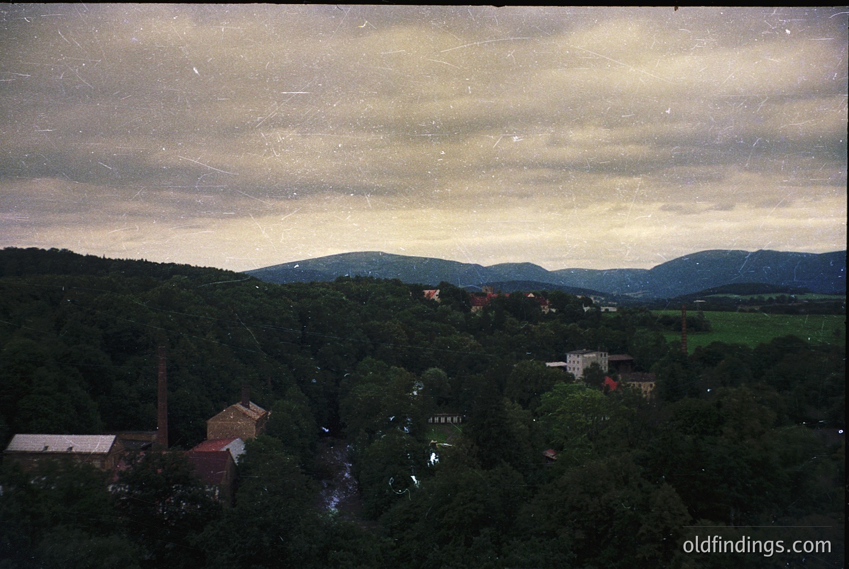 Vintage aerial view of a forested valley with industrial buildings and scattered structures. Dense tree cover dominates the landscape, with a prominent brick chimney and red-roofed factory in the foreground. Rolling hills and distant mountains frame the horizon under a cloudy sky. Likely Eastern European industrial region, mid-20th century.