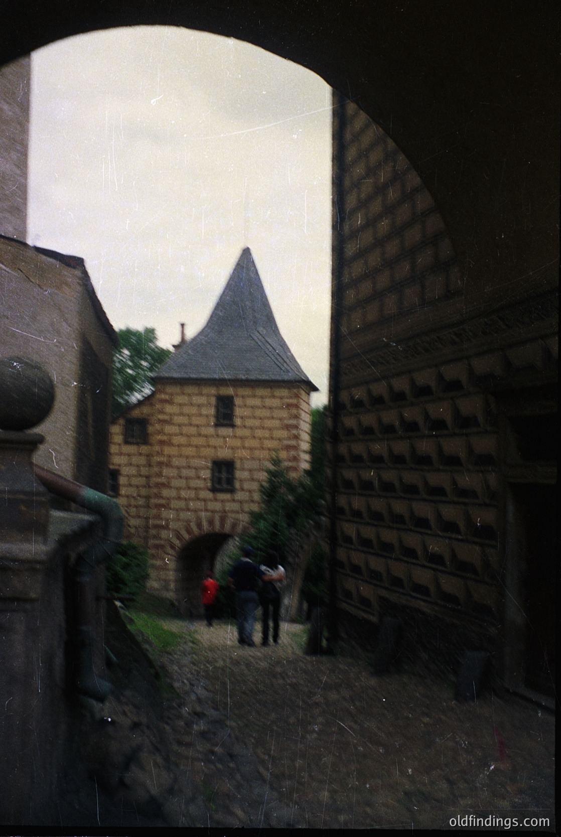 Medieval-style courtyard framed by arched stone walls, featuring a small tower with conical roof and brickwork. Two figures in modern clothing walking toward an arched passageway. Likely European castle or fortress, 12th–16th century.