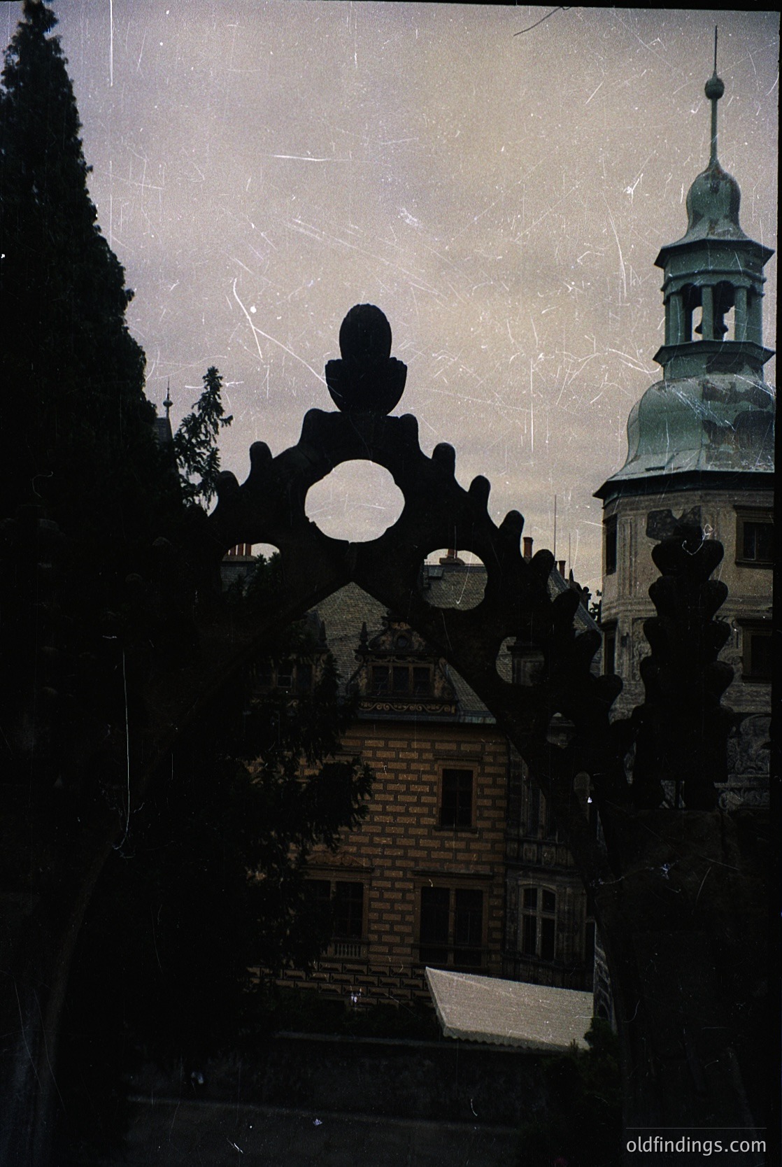 Vintage architectural silhouette framed by ornate wrought-iron balcony railing. Gothic Revival-style building with pointed tower and decorative stonework, likely 19th-century European. Overcast sky enhances dramatic contrast.