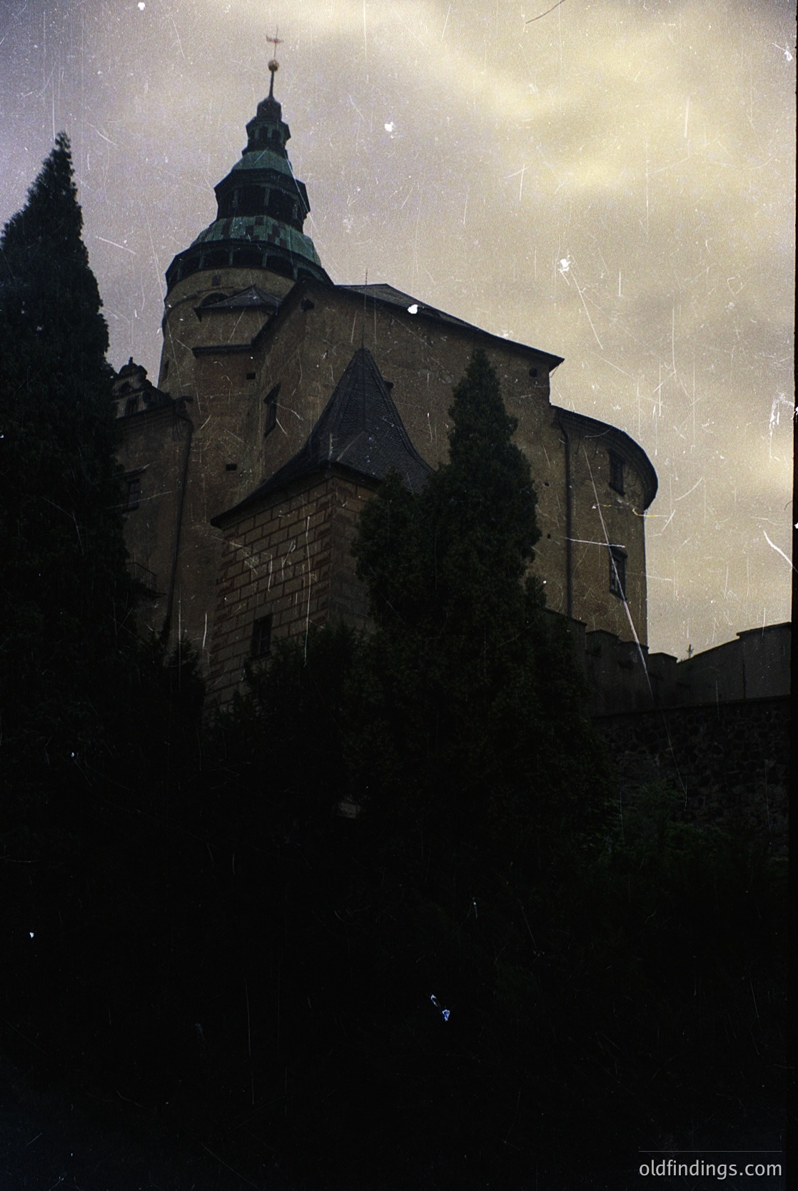 Historic stone church with Gothic Revival architecture—distinctive steep gables, central tower, and green patinated spire. Overgrown foliage frames the lower facade. Vintage sepia tone suggests mid-20th century photography. Potential European setting.