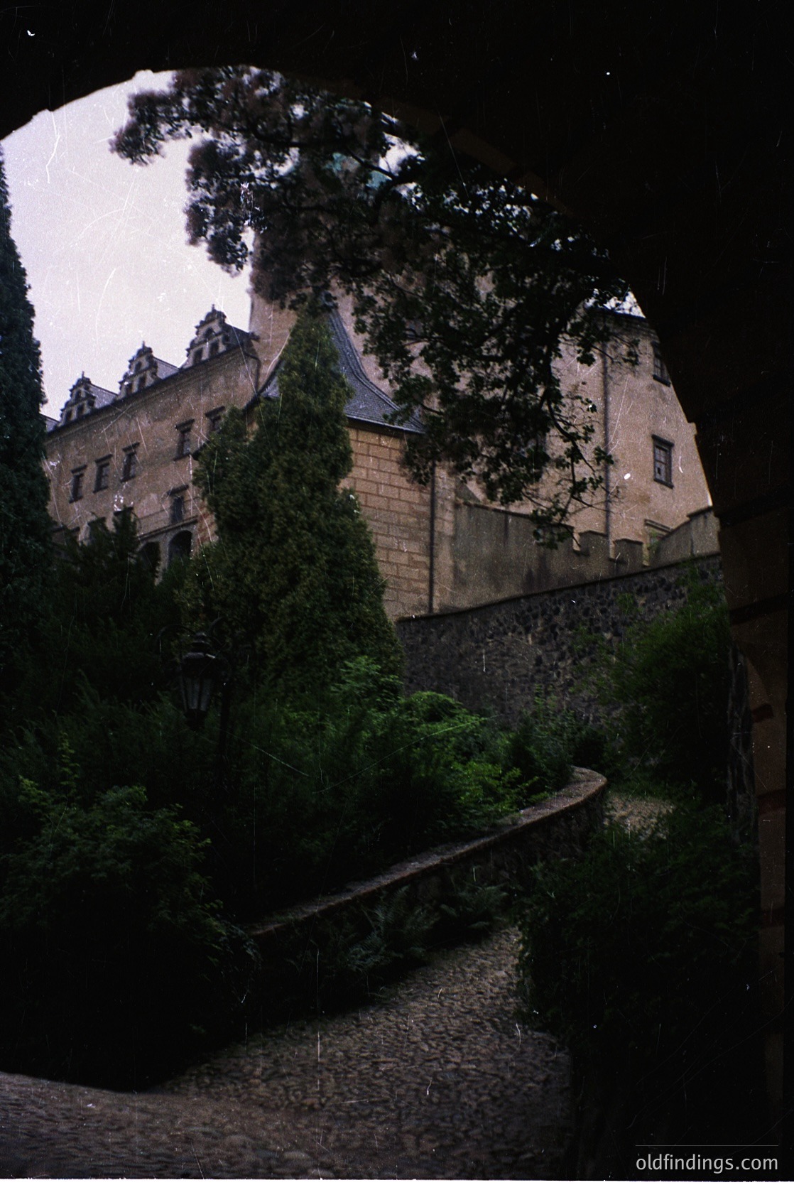 Historic stone castle framed by dense foliage, illuminated by soft evening light. Gothic-style architecture with steep gables and ivy-covered walls. Cobblestone pathway curves beside a shallow stream, accented by vintage-style lanterns. Likely European, possibly or early .