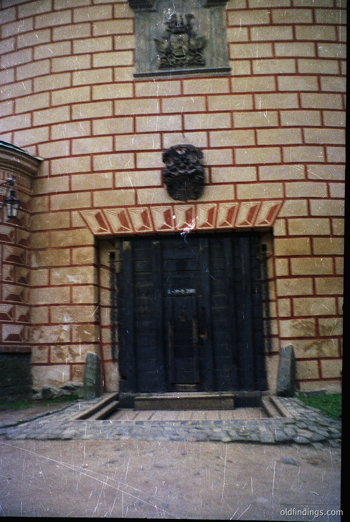 Historic brick entrance with ornate wrought-iron double doors framed by red-and-white striped stonework. Decorative stone carvings above the door and atop the arch. Cobblestone pathway and weathered stone steps lead to the entrance, suggesting European architectural heritage.