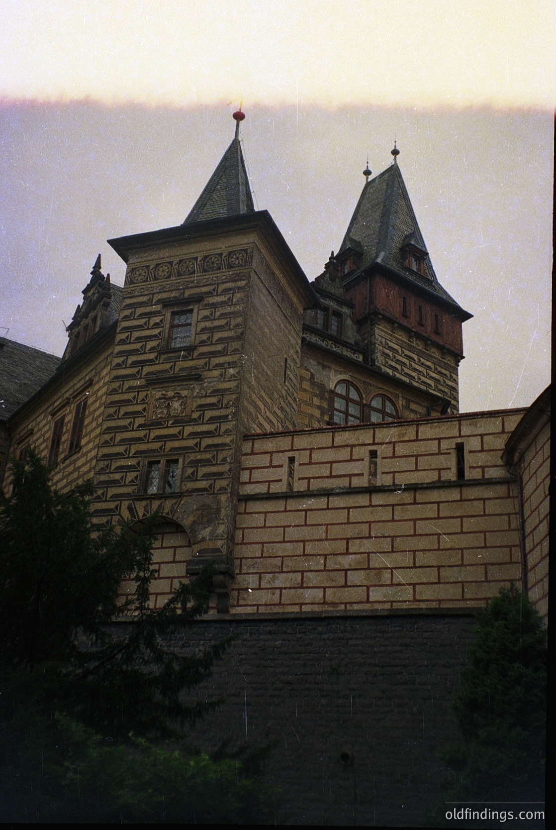 Neoclassical stone fortress with twin conical towers and crenellated walls, likely European. Ornate stonework and arched windows suggest 19th-century military architecture. Dramatic evening light enhances texture.