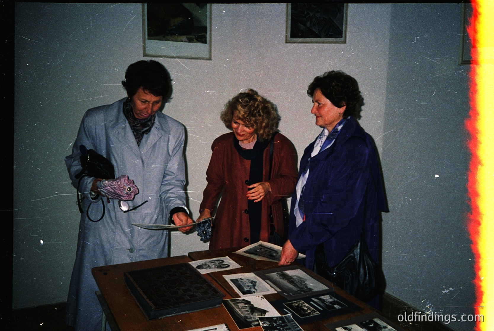 Three individuals examine framed photographs on a display table in a dimly lit indoor setting, likely a museum or gallery. The women’s 1970s–1980s attire includes coats, scarves, and shoulder bags. The table holds black-and-white and color images, possibly historical or artistic.