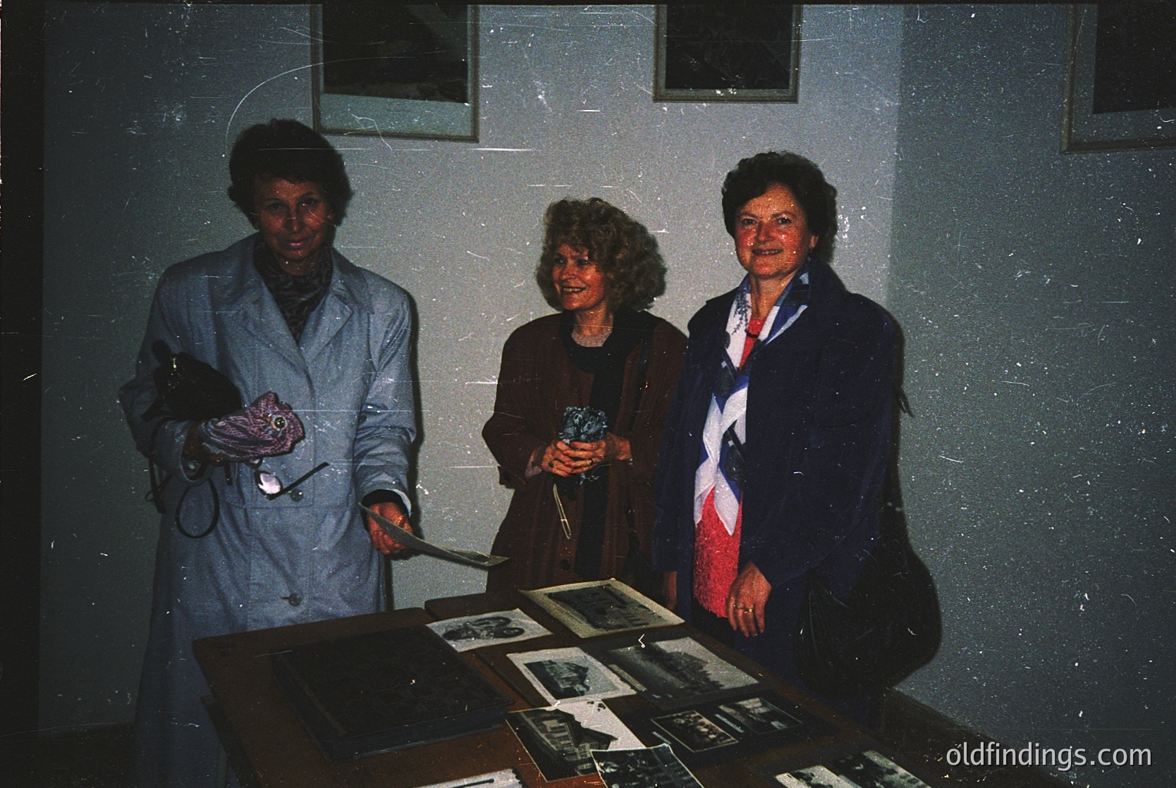 Three women pose indoors, likely in a medical or institutional setting from the 1970s–1980s. Left: nurse in white lab coat holding stethoscope. Center: woman in dark dress with curly hair. Right: woman in blazer with red-and-white striped tie. Display table behind them features framed black-and-white photographs. Dim lighting and textured wall suggest a formal or commemorative event.