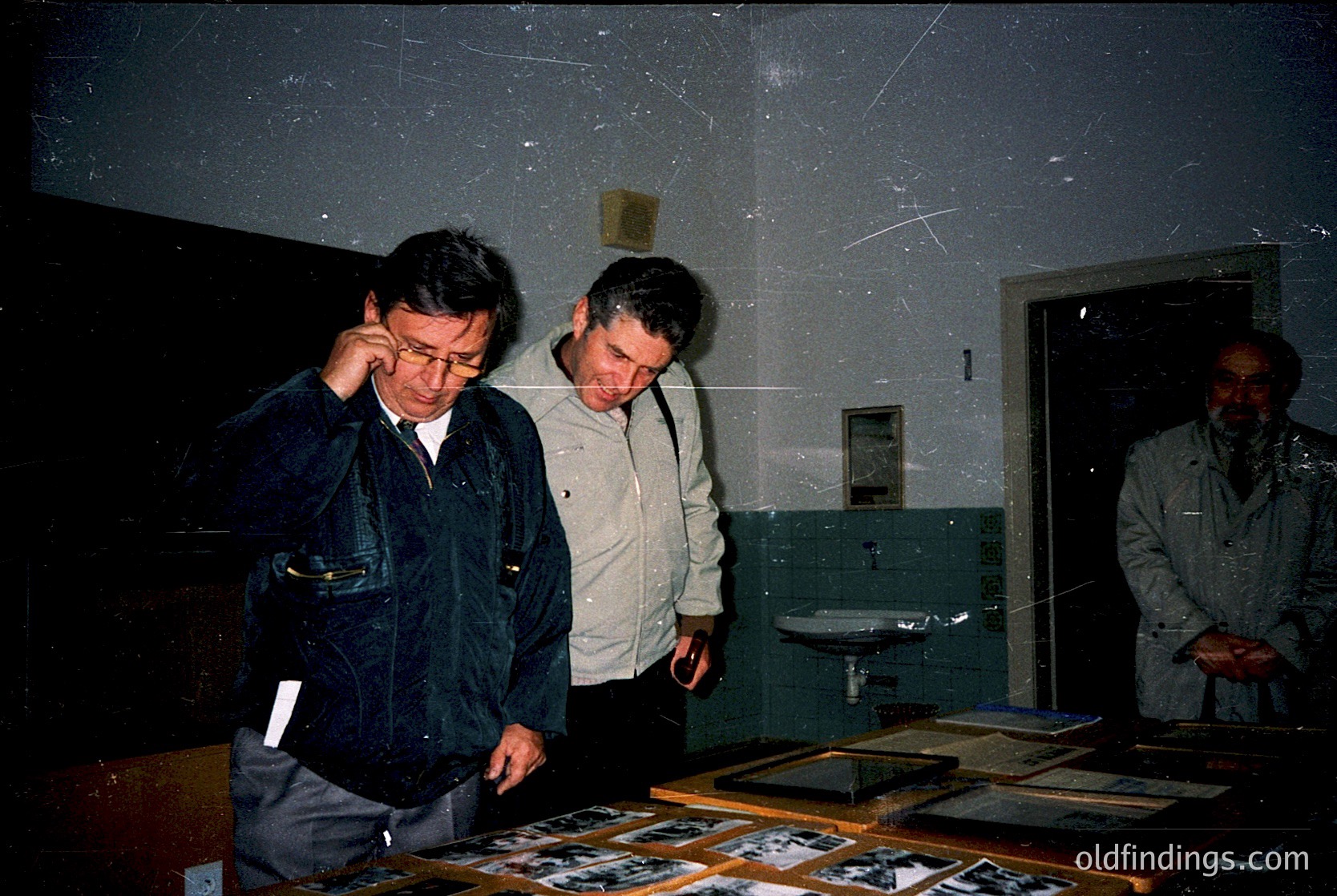 Three men examine framed black-and-white photographs in a dimly lit room, likely a museum or archive. The man on the left wears a dark jacket and glasses, while the others in neutral tones. Shelves display additional framed images. The setting suggests a 1980s–1990s Eastern Bloc or Soviet-era institution.