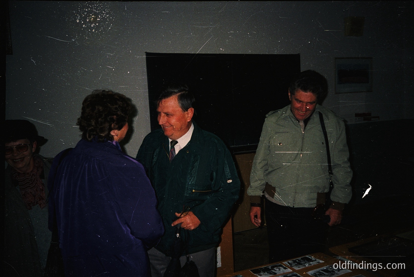 Indoor gathering featuring three men in 1970s-era attire—dark blazers, collared shirts, and shoulder bags—engaged in conversation. Dim lighting reveals a table with printed materials, likely documents or photos. Background shows a framed poster and a textured wall. Style suggests a formal or professional setting, possibly a conference or meeting.