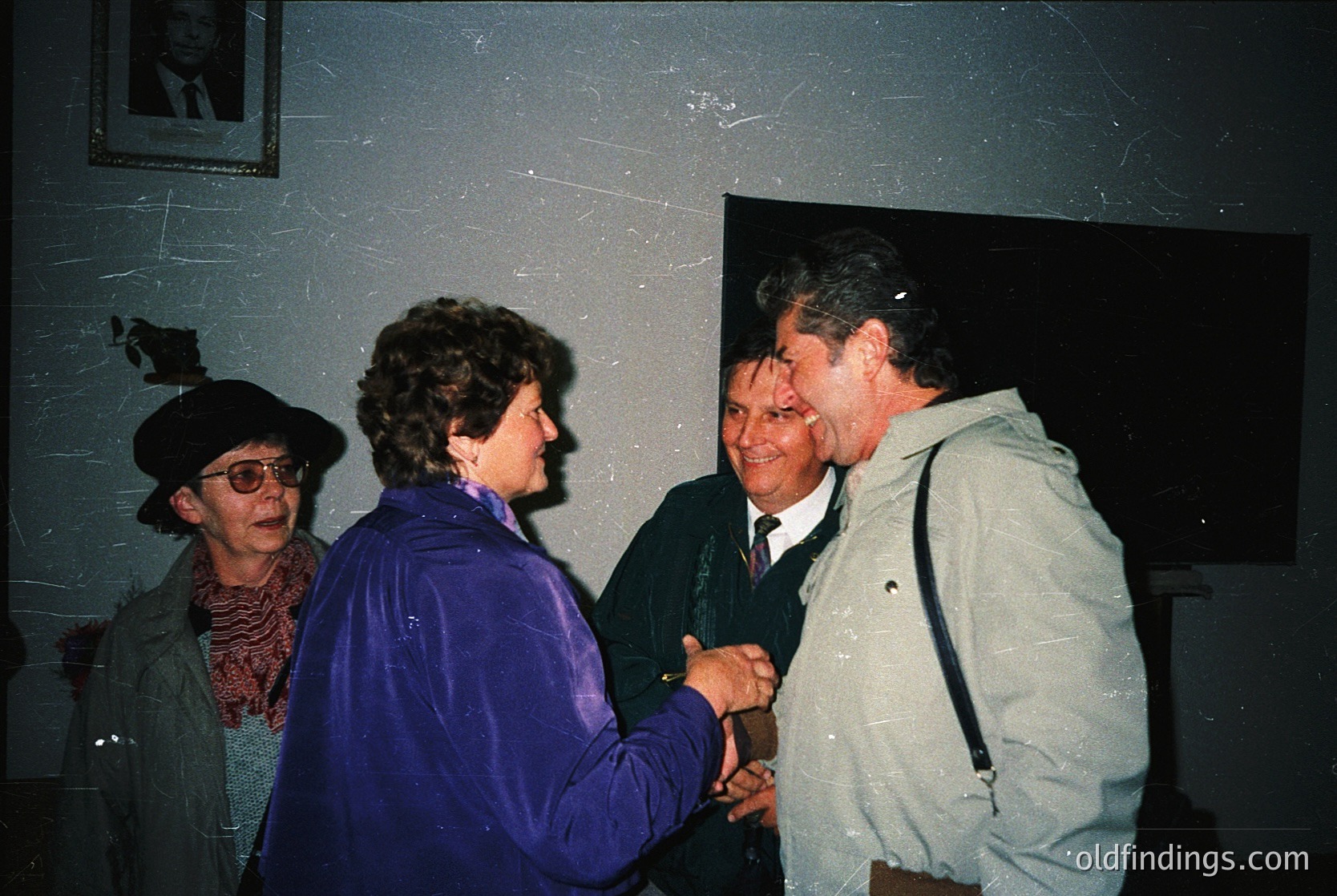 Indoor gathering featuring four adults in mid-1990s attire. A woman in a purple jacket shakes hands with a man in a light jacket and backpack, while another woman in a black hat and scarf observes. A fourth man in a dark suit stands nearby. Plain walls and a framed portrait suggest a formal or semi-formal event.