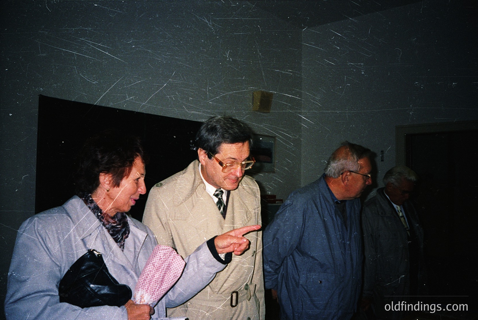 Four adults in formal attire—men in blazers, women in coats—gather indoors, likely at a conference or exhibition. The central man gestures toward an unseen display, while others observe. Dim lighting and patterned wallpaper suggest a mid-20th-century venue. Style hints at 1970s–1980s professional or academic setting.