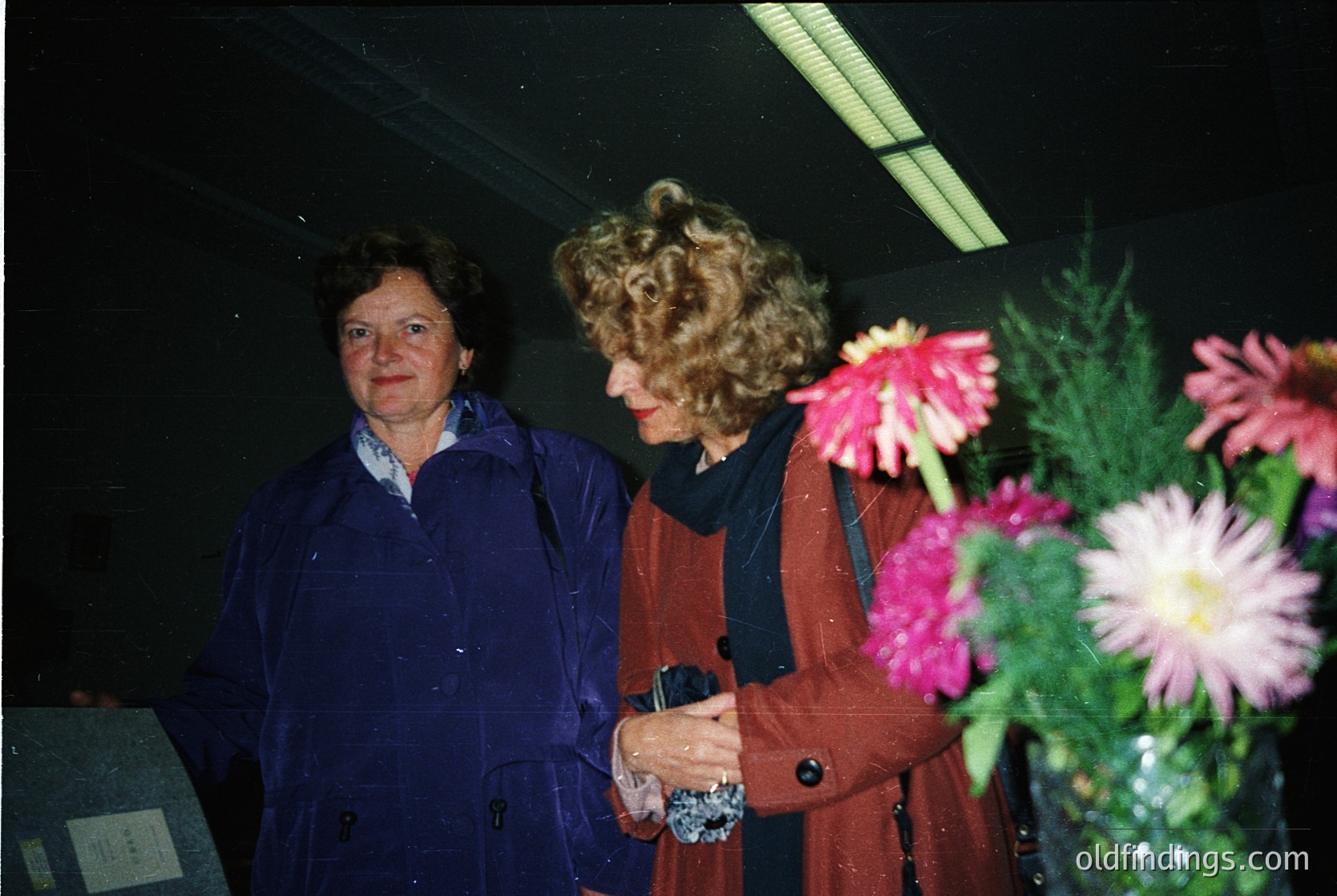 Two women in 1980s/90s attire pose indoors near vibrant flowers. The woman on left wears a dark blue coat; the right, a maroon blazer with curly hair. Fluorescent lighting and floral arrangement suggest an event or office setting.