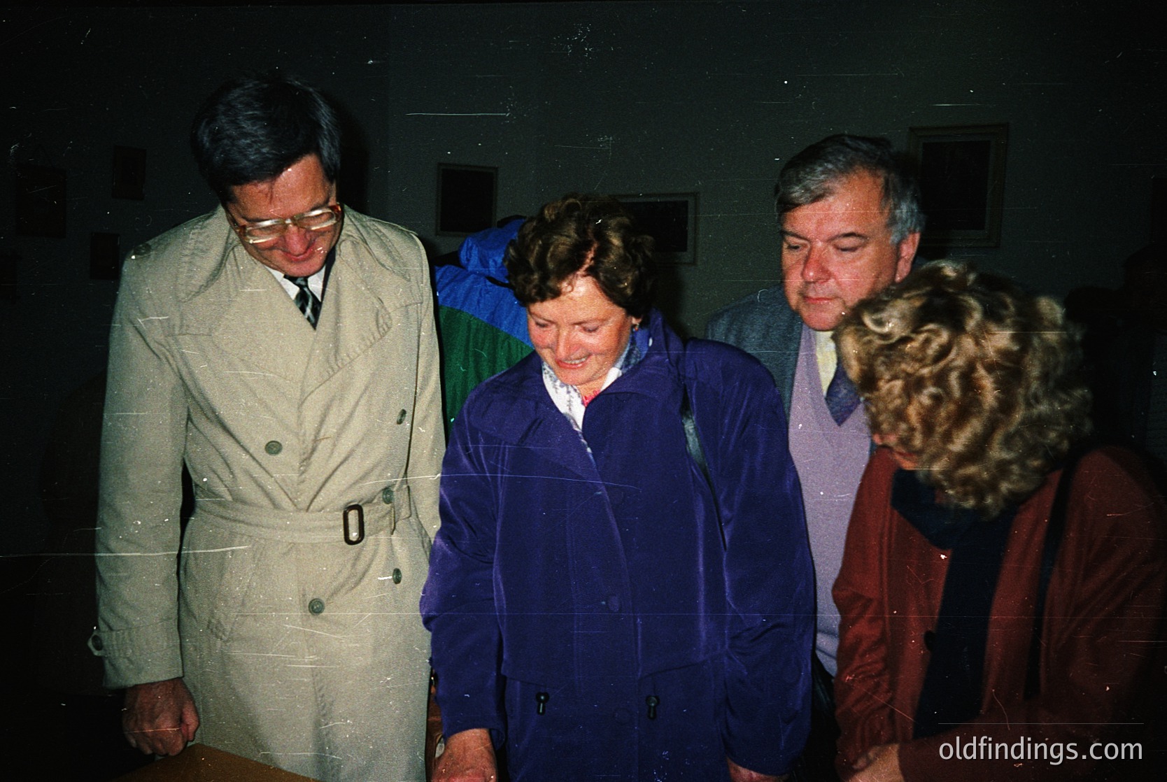 Four individuals in 1970s-era attire—men in blazers, women in raincoats—pose indoors under dim lighting. The man on the left wears glasses and a light-colored trench coat; the woman in blue stands centrally, smiling. The background suggests a formal or institutional setting, possibly a conference or office.
