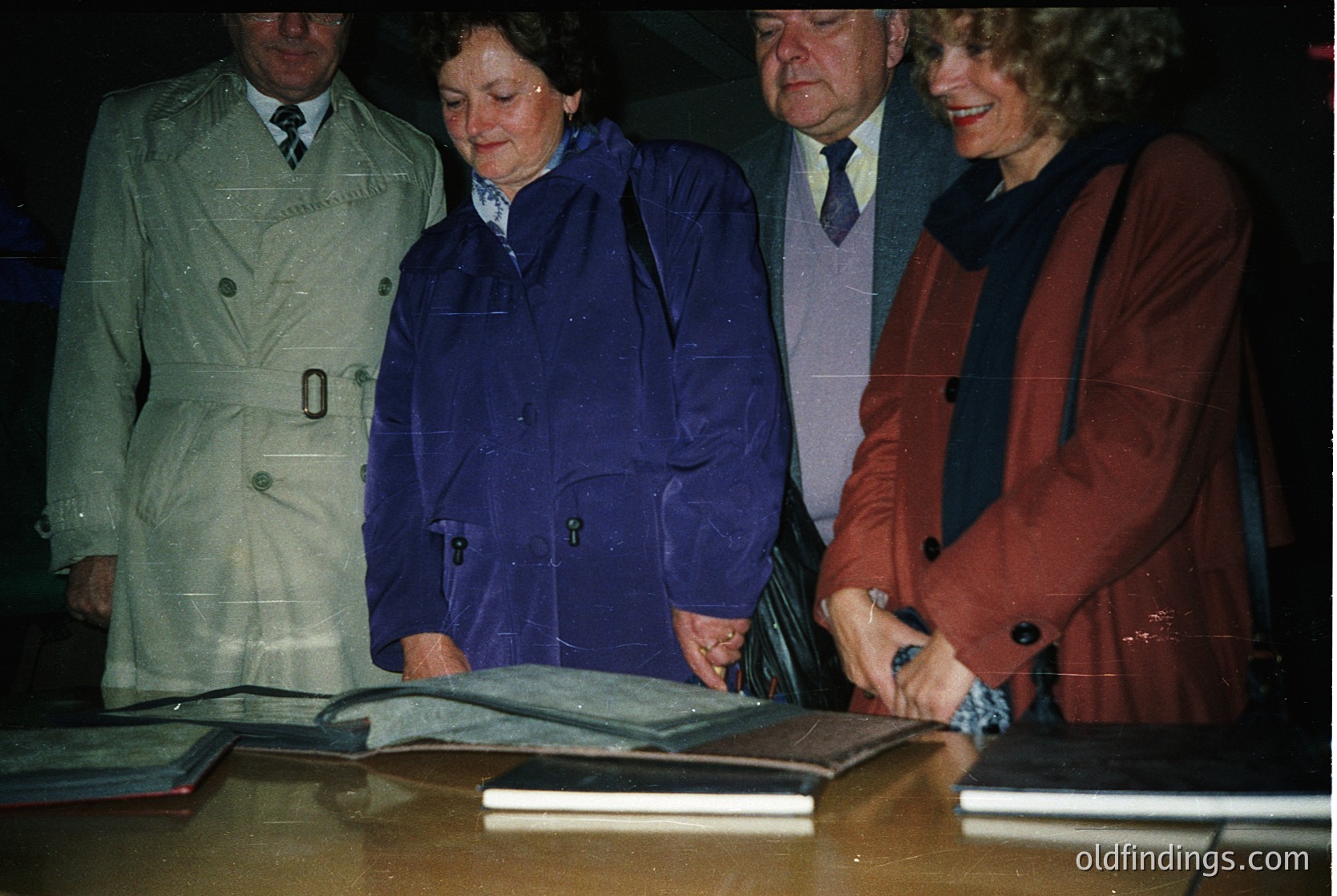 Four individuals in formal attire inspect a large, open book on a table, likely at a ceremonial signing event. Men wear suits with ties; women in coats and scarves. Indoor setting with muted lighting, suggesting a formal occasion.