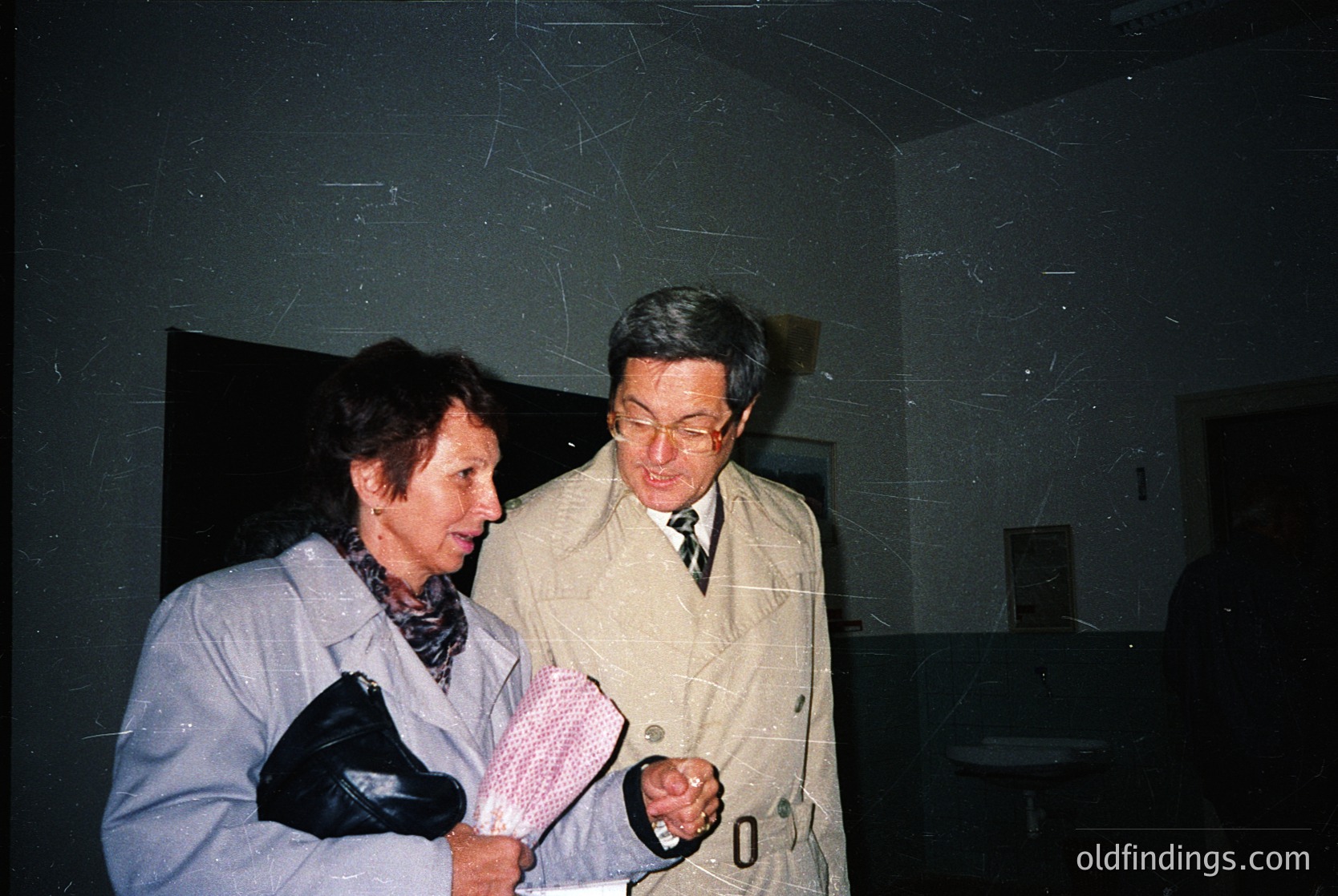 Two individuals in formal attire converse indoors, likely in a public or institutional setting. The woman wears a light gray coat and holds a white handkerchief; the man in a beige trench coat with a watch and tie. The dim lighting and aged wall texture suggest a mid-20th century setting ( ). The scene evokes historical or professional documentation.