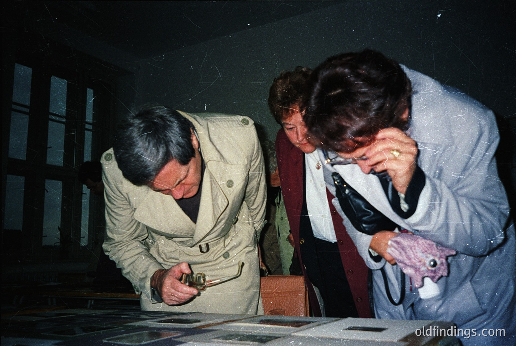 Three individuals examine vintage photographs in a dimly lit room, likely a museum or archive. The man in the center wears a beige coat with a visible "U" emblem, while the woman on the right holds a pink object and wears a white lab coat. The table displays framed black-and-white images, suggesting a historical or cultural exhibit. The setting appears to be mid-20th century, possibly Eastern Europe. [Historic photo exhibit with three figures examining vintage prints in a museum setting, mid-20th century ]