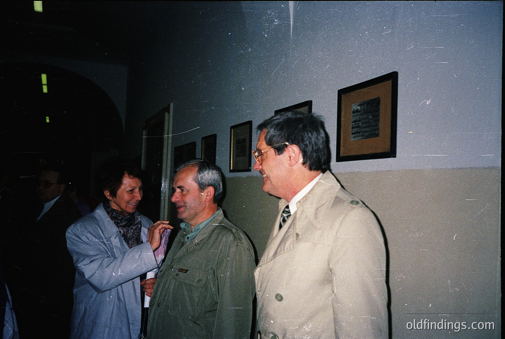 Indoor gathering featuring three men and a woman in formal attire, likely late 20th century. The man in olive drab uniform (likely military) stands beside two men in suits—one in beige, one in dark—with a woman in a light blue coat. Framed plaques on the wall suggest an institutional or ceremonial setting. Low-key lighting and muted colors indicate a formal event.