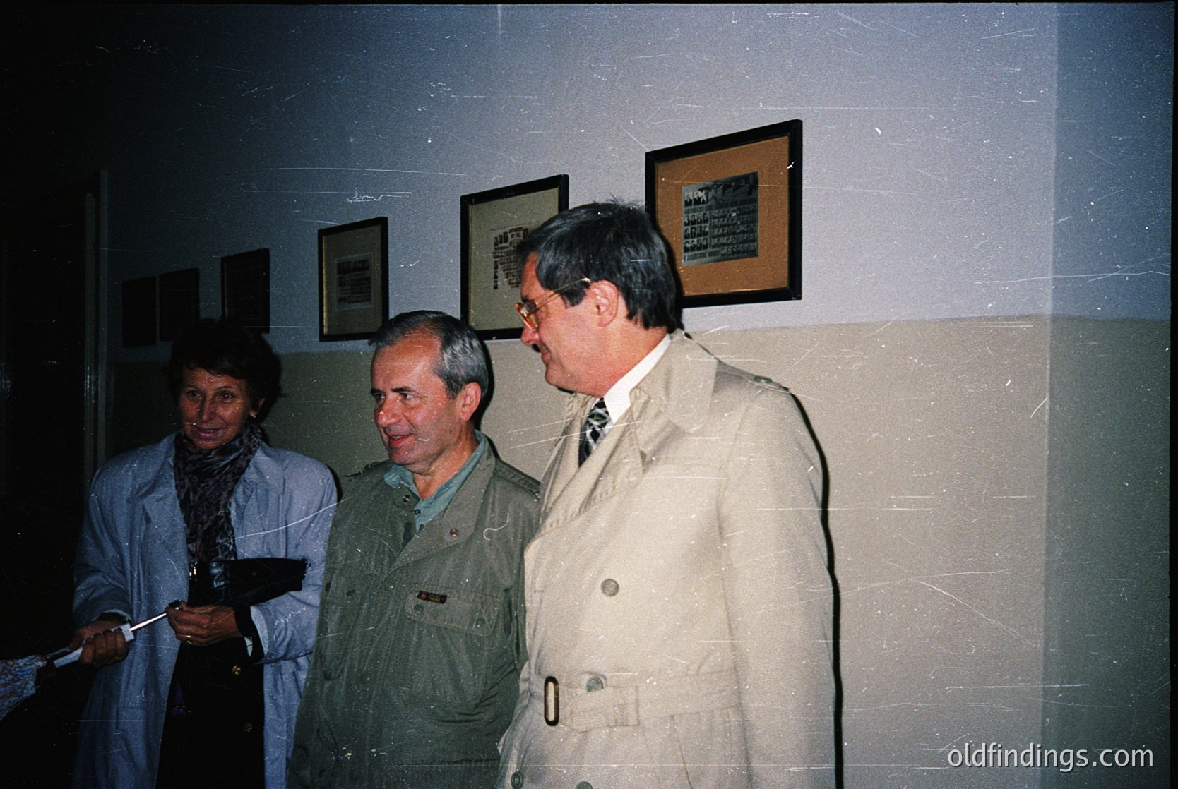 Three individuals in formal attire pose indoors, likely at a 1980s–1990s event. The man in the center wears a green jacket with a patch, while the man on the right dons a beige trench coat. The woman on the left holds a clipboard and wears a dark coat with a scarf. Plain wall with framed plaques suggests an institutional or official setting.