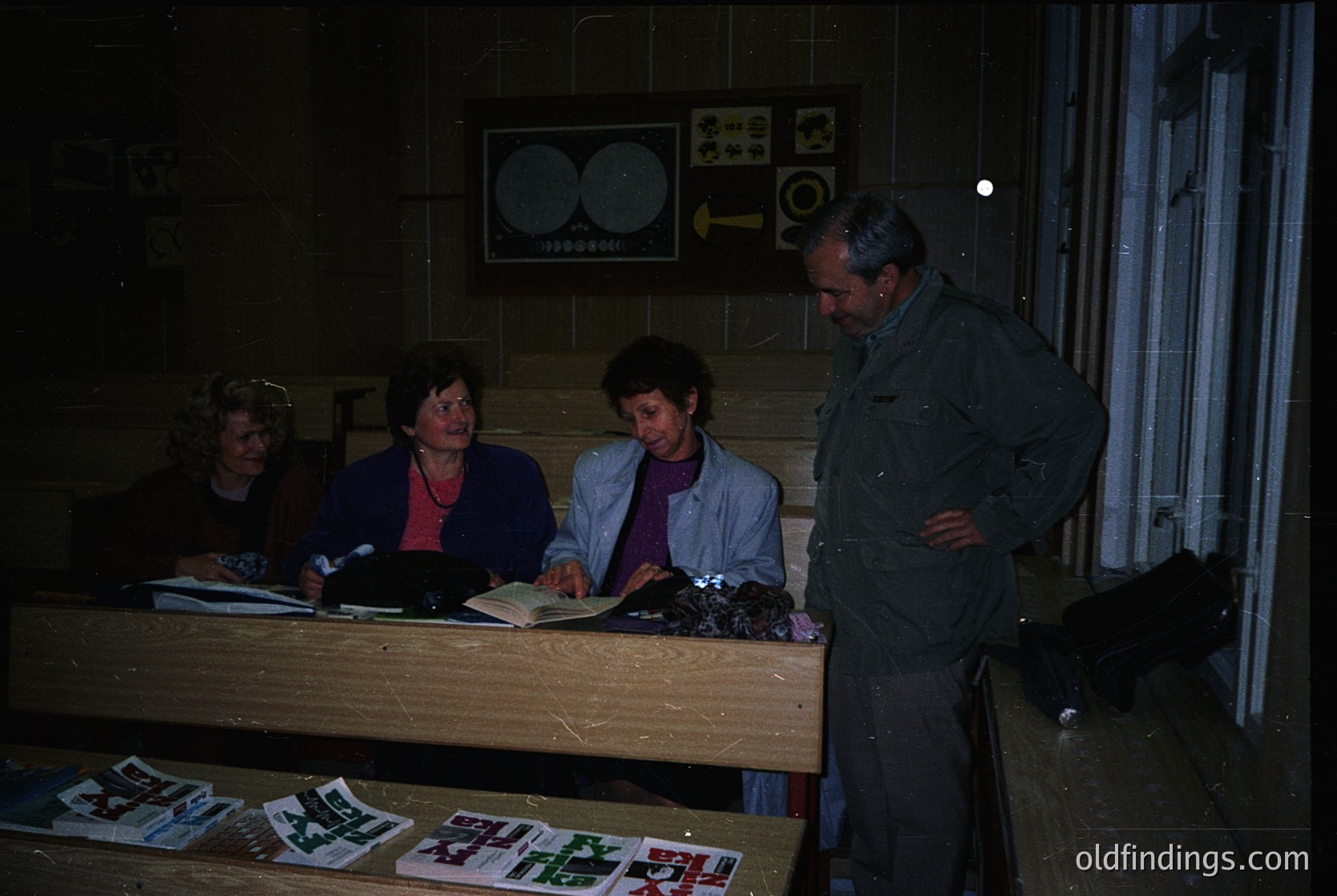 Four individuals in a dimly lit indoor setting, likely a 1970s-80s office or archive. Two women seated at a wooden desk reviewing documents, one man standing with hands on hips, another examining a book. Posters with celestial maps and directional arrows on wall. Stacks of printed materials on desk.