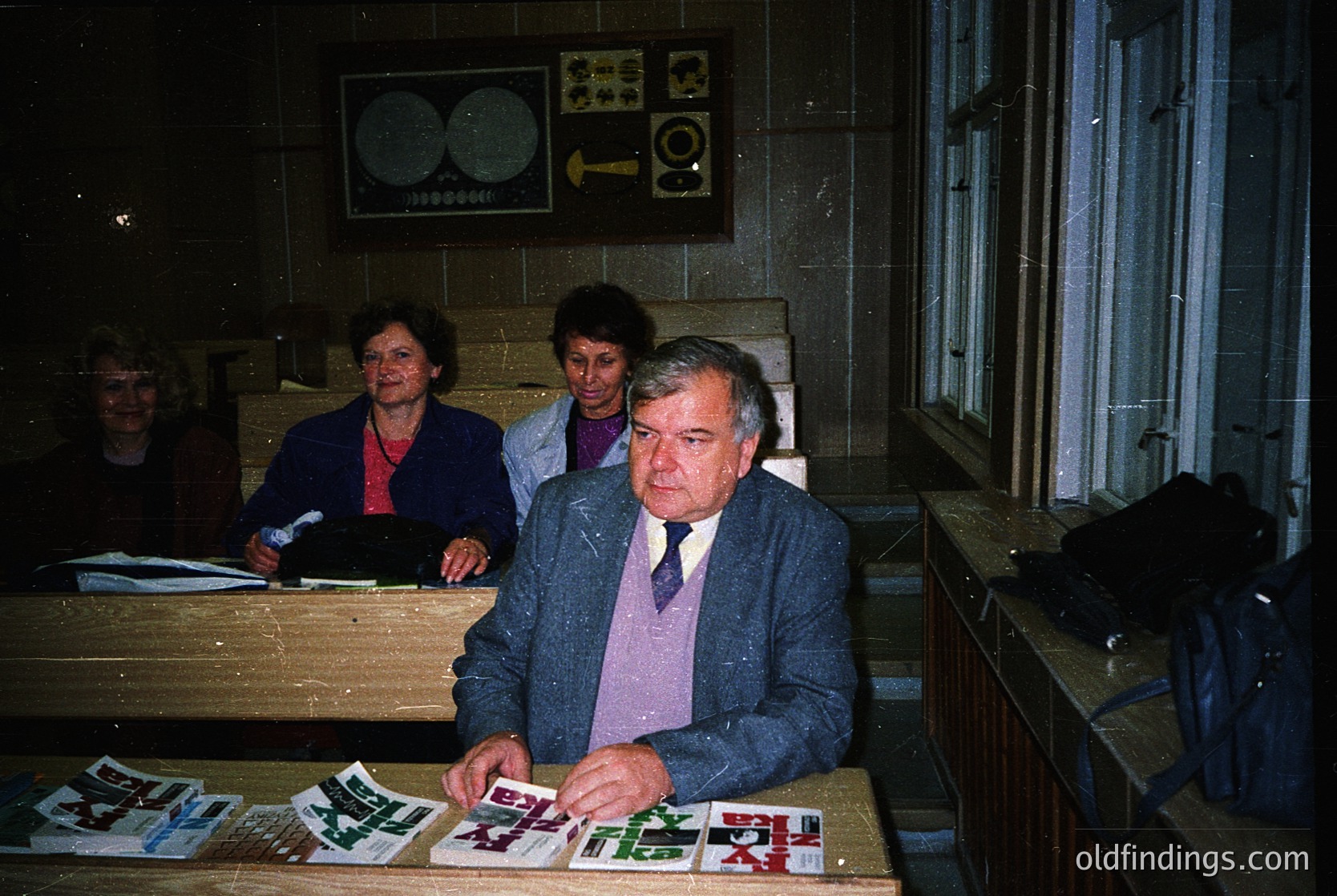 Indoor office setting from the 1970s-1980s, featuring a man in a suit and tie reviewing printed materials on a wooden desk. Behind him, three women in business attire stand near a wall-mounted clock and directional sign. Shelving with files and a coat rack occupy the right side. Warm lighting and dated office decor suggest a mid-century administrative environment.