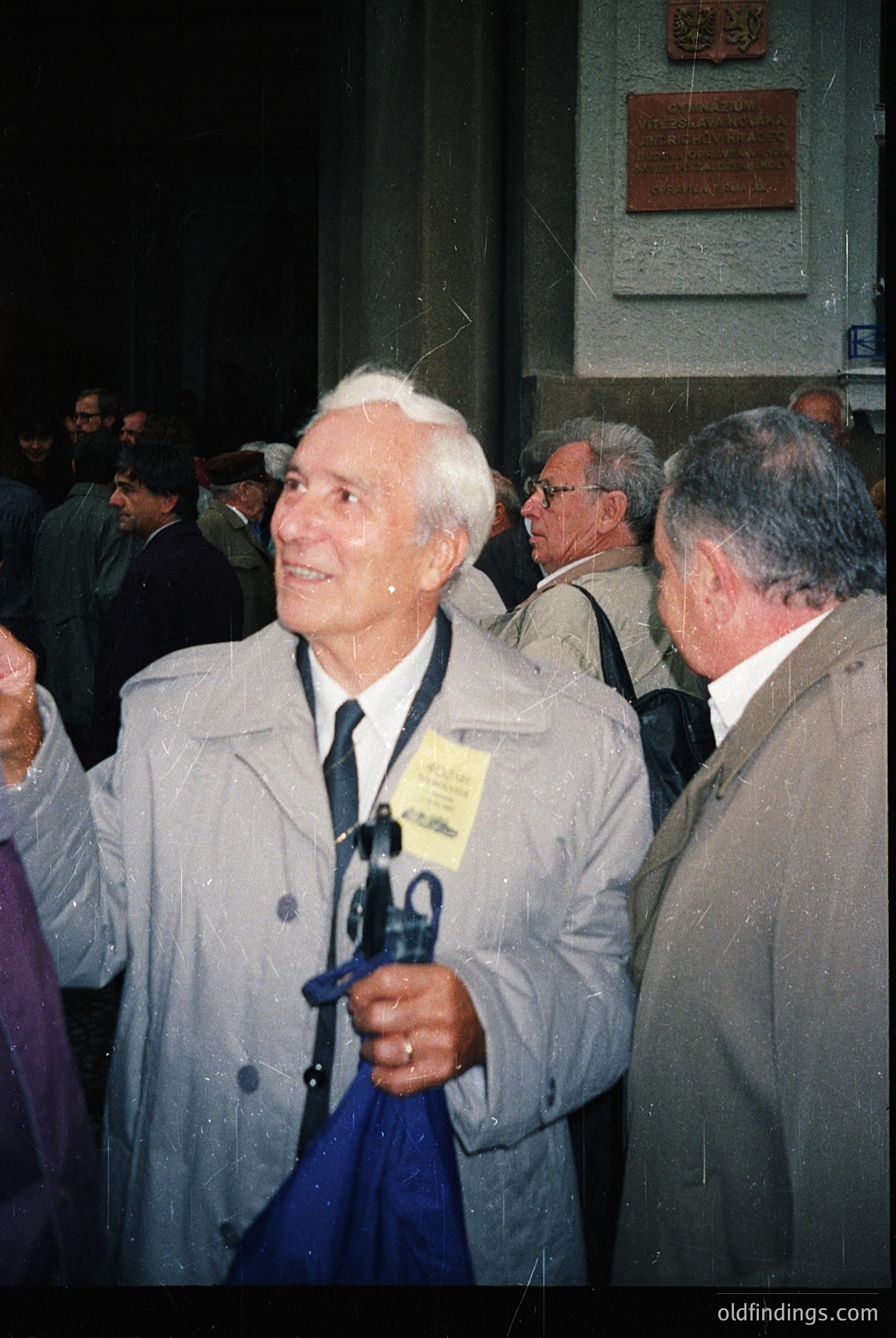 Mid-20th century formal event with elderly man in light coat, holding a blue umbrella and lanyard badge. Crowded indoor setting with classical architecture (plaque, columns). Likely Eastern Bloc era (1960s–1980s).