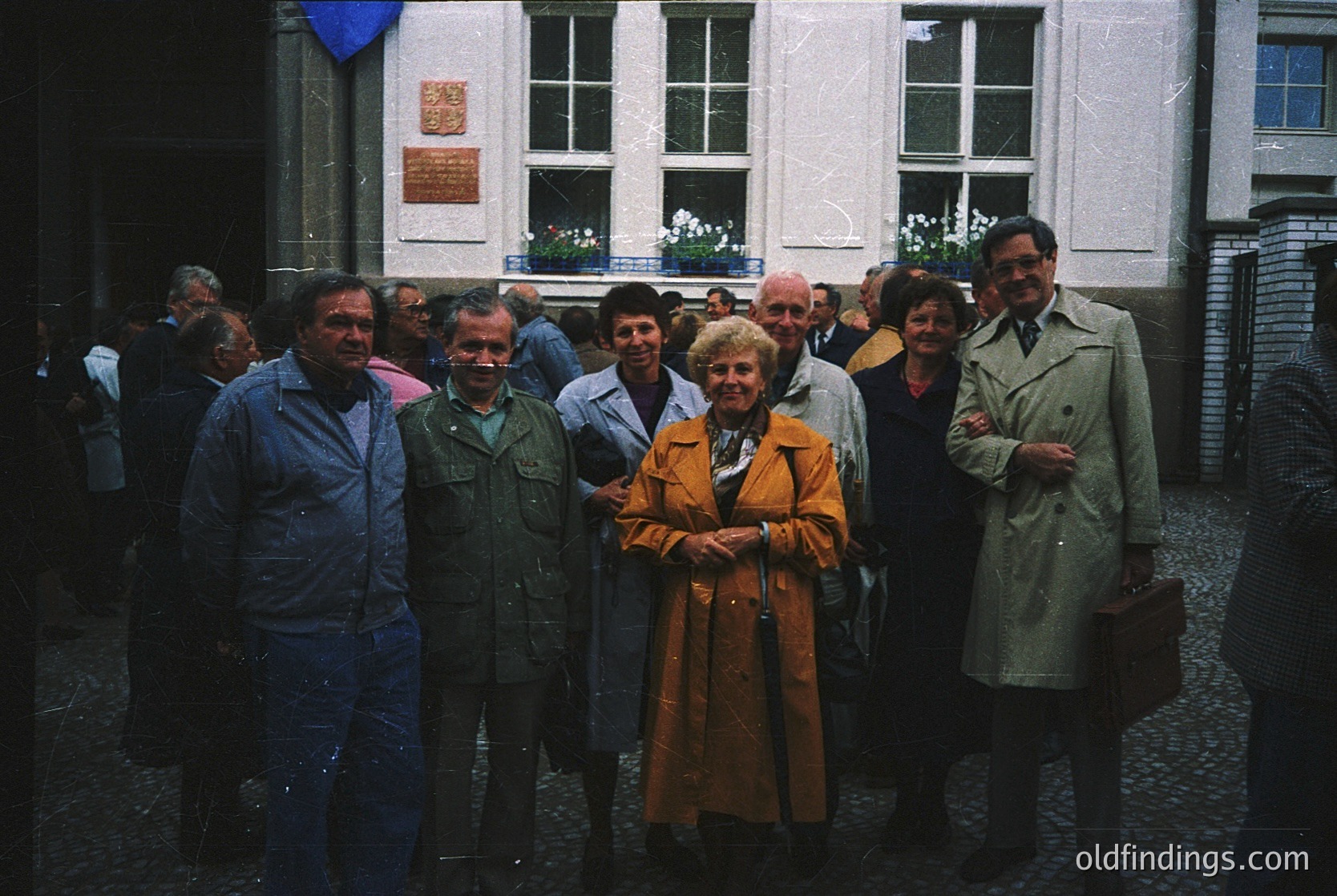 Group portrait in a European courtyard, likely 1970s–1980s. Eight individuals pose in vintage coats, scarves, and glasses, standing on cobblestone pavement. A plaque on the building’s facade suggests institutional or governmental significance. Warm sepia tones enhance nostalgic atmosphere.