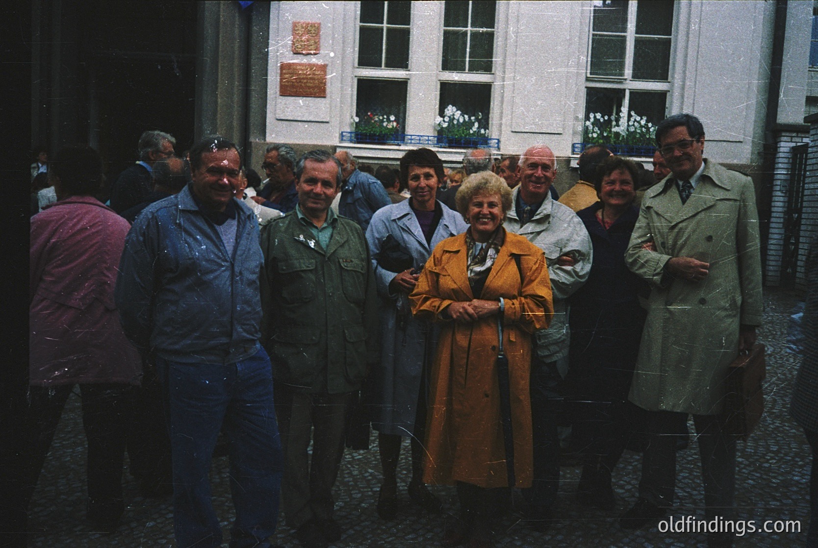 Group of 10 adults posing outdoors in 1970s European urban setting. Central woman in mustard-colored dress, surrounded by men in jackets and women in coats. Cobblestone street, brick buildings with large windows and potted plants. Formal, candid group portrait style.