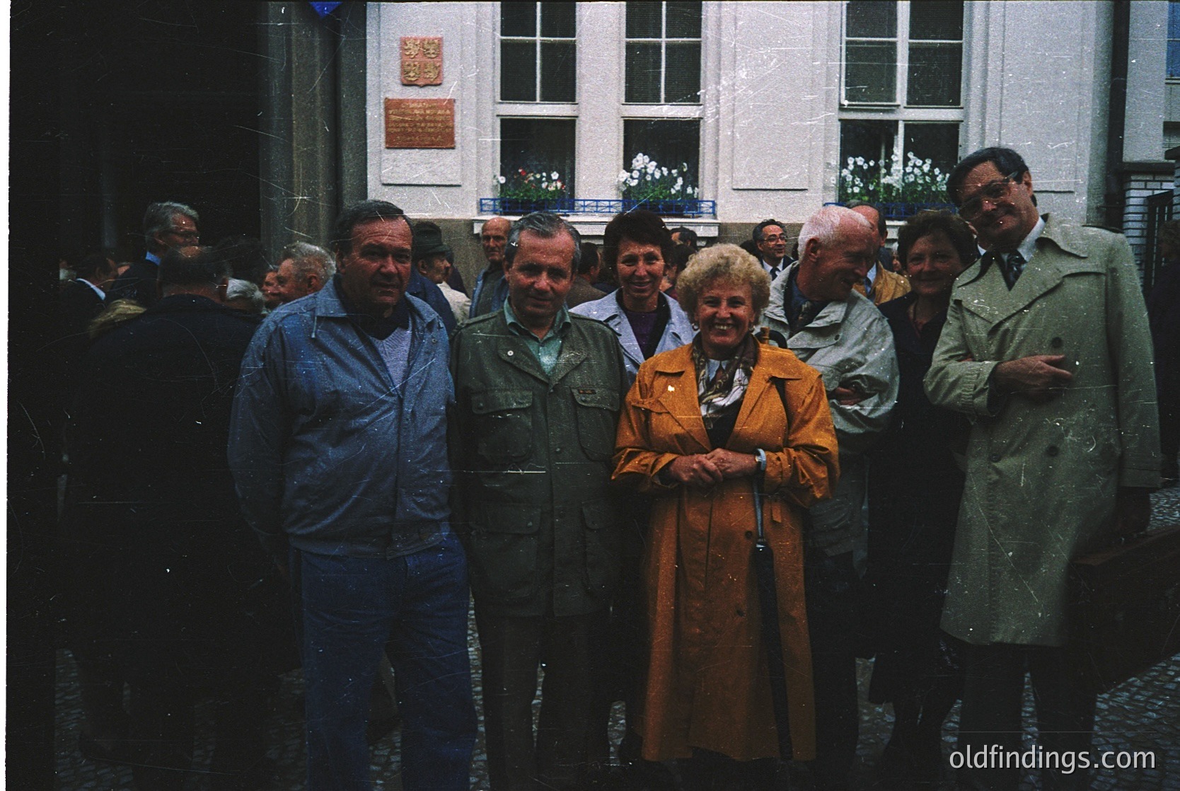 Group of seven adults posing in front of a European courtyard building, likely 1970s–1980s. Men wear jackets, ties, and overcoats; women in coats and blouses. Cobblestone ground and brick walls with a plaque and large windows. Formal, possibly professional or cultural event.