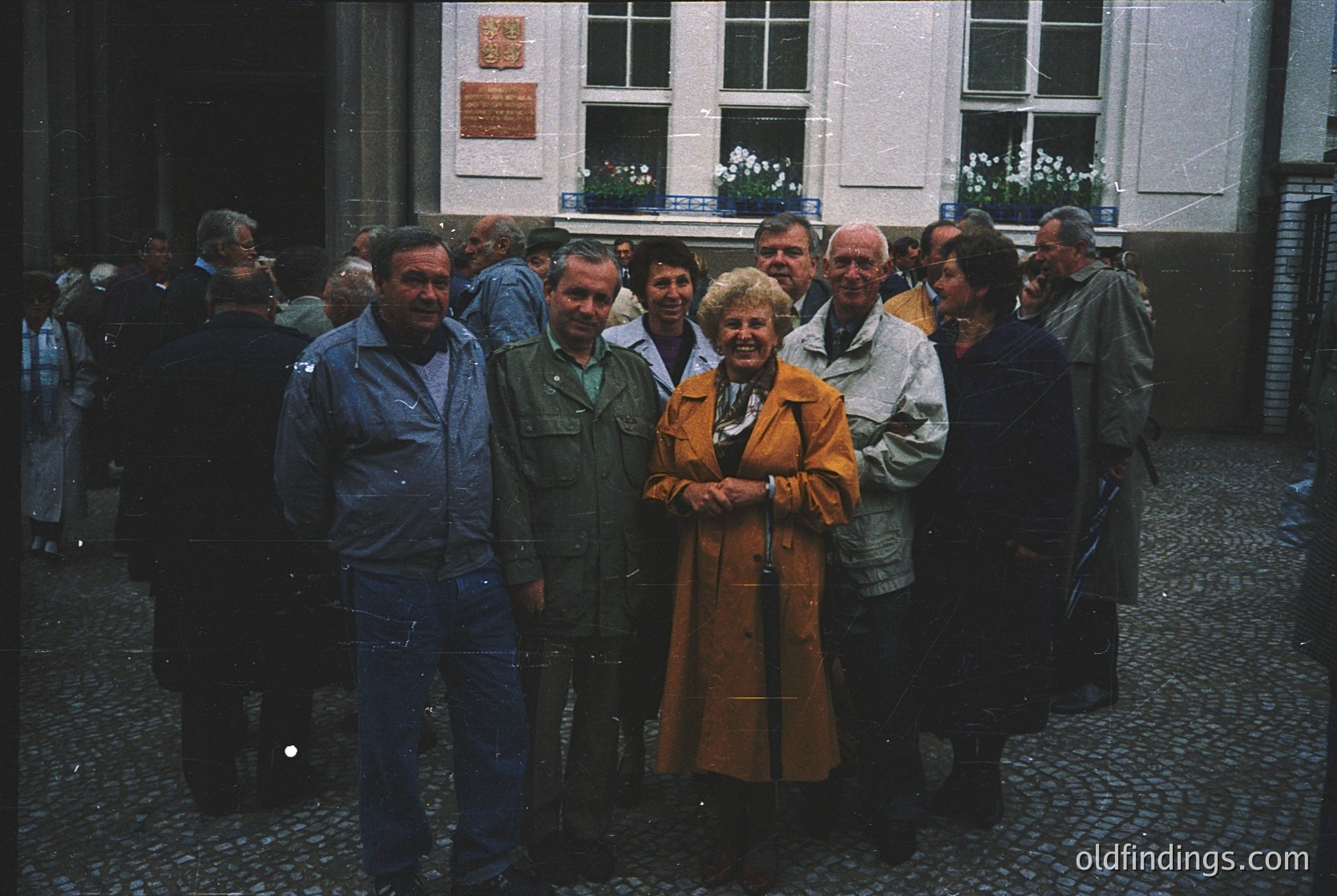 Group photo in a European courtyard, likely 1970s–1980s. Cobblestone ground and brick buildings with white-framed windows frame the scene. Central woman in a mustard coat stands with a cane; others in jackets and caps pose casually. Warm sepia tone suggests vintage film.