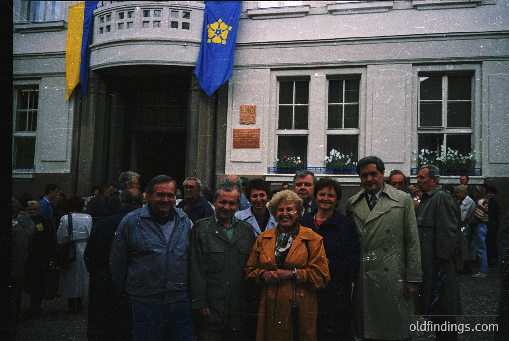 Neatly arranged group photo in front of a Soviet-era building, featuring a yellow sunburst emblem on a blue flag. Eight men and women in 1970s attire pose formally, with some wearing jackets and ties. The building’s classical facade includes rectangular windows and a plaque. Crowd visible in background.